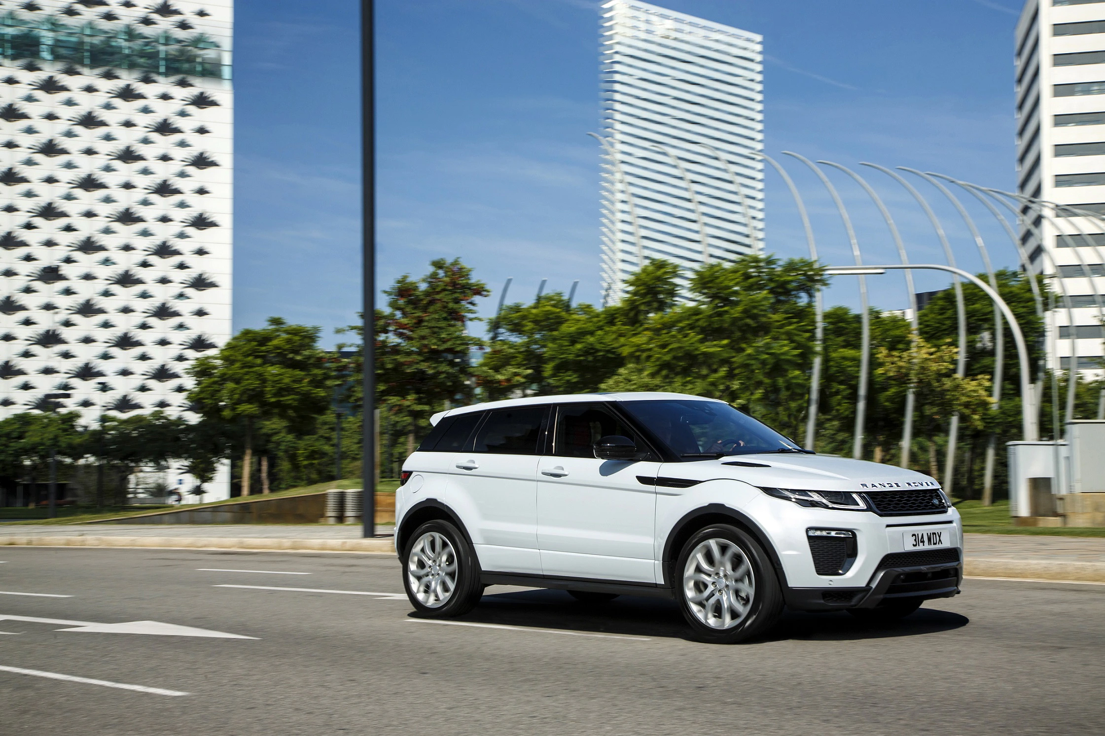 Ein weißer SUV fährt auf einer städtischen Straße. Im Hintergrund sind moderne Gebäude und ein blauer Himmel sichtbar.