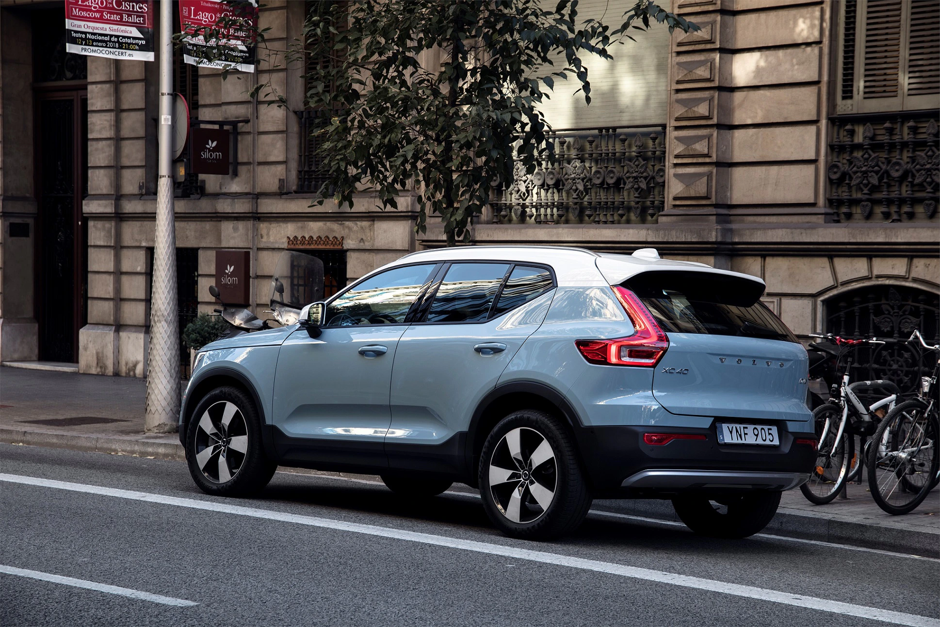 Blauer SUV parkt auf einer Stadtstraße neben einem Gebäude mit Fenstern und Fahrrädern. Im Hintergrund sind Schilder und Bäume zu sehen.