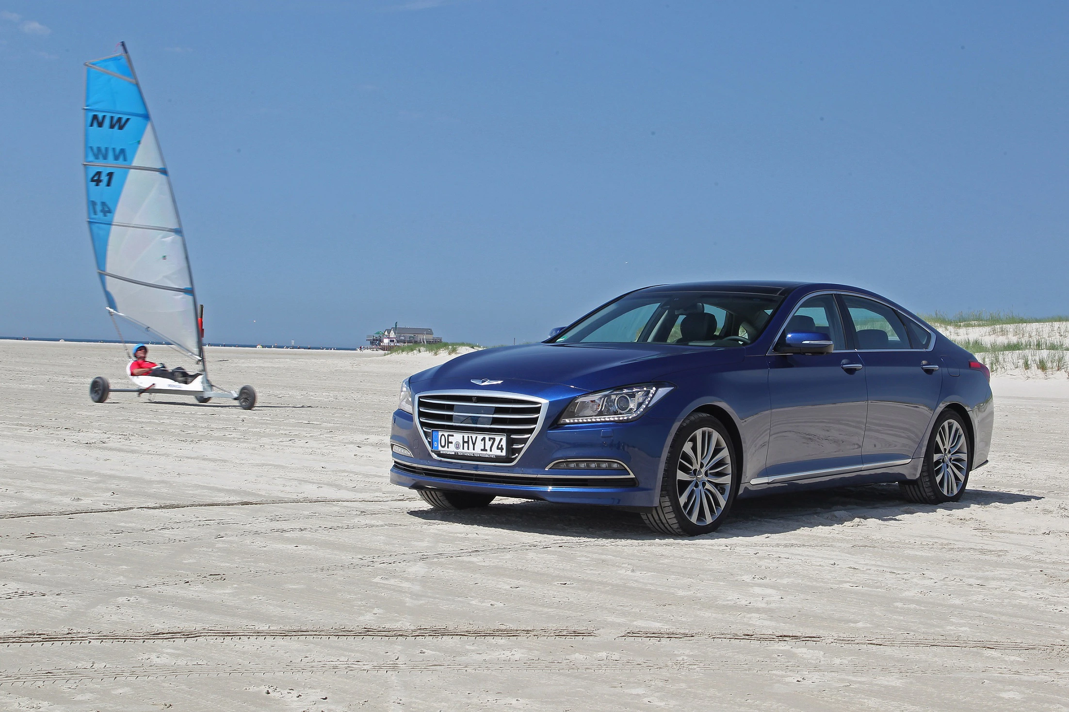 Blaues Auto parkt am Strand, während ein Strandsegler im Hintergrund vorbeifährt. Klarer Himmel und Sanddünen im Hintergrund.
