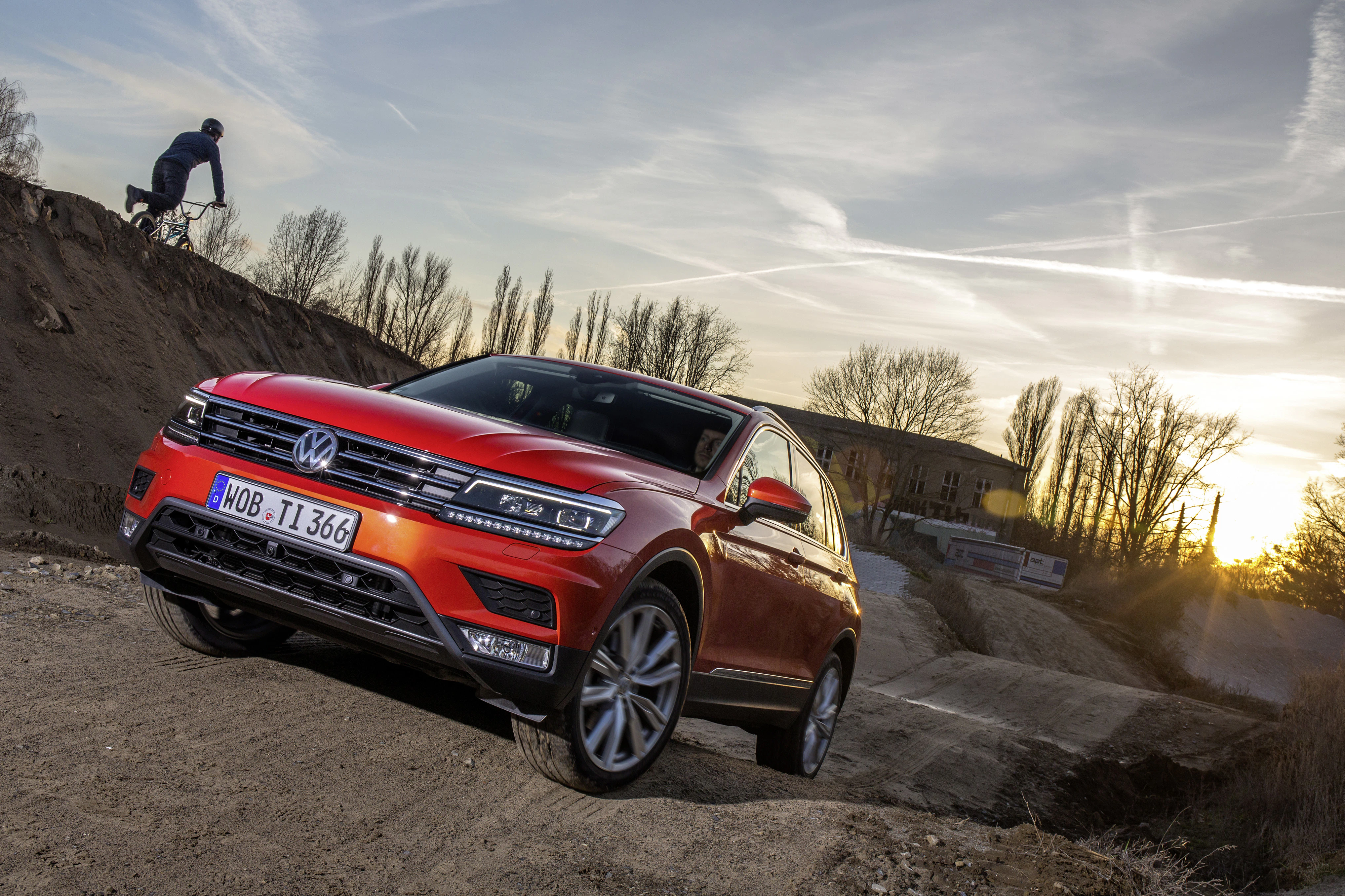 Roter Volkswagen SUV fährt eine sandige Piste hinauf bei Sonnenuntergang. Ein Radfahrer ist im Hintergrund auf einem Hügel zu sehen.