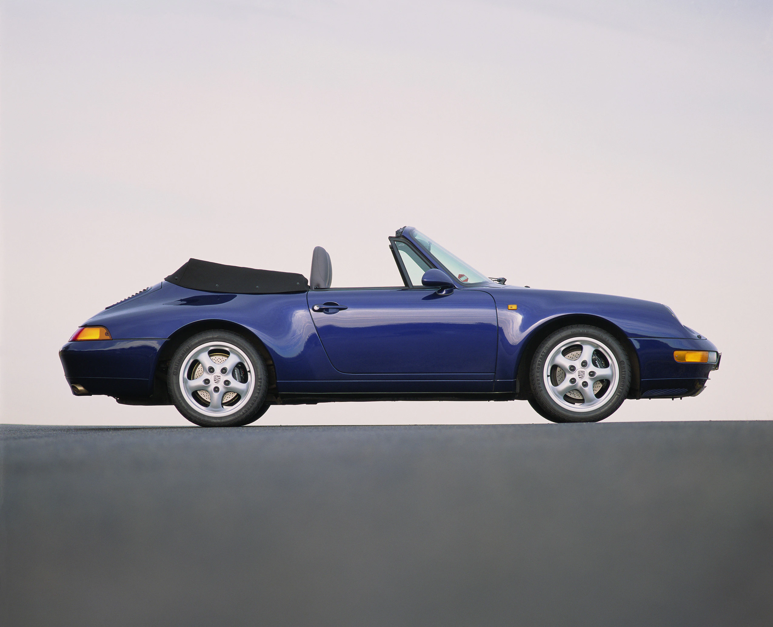 Blaues Cabrio mit geöffnetem Verdeck auf freier Fläche, seitliche Ansicht. Elegantes Design, klassische Linien und sportliche Räder.