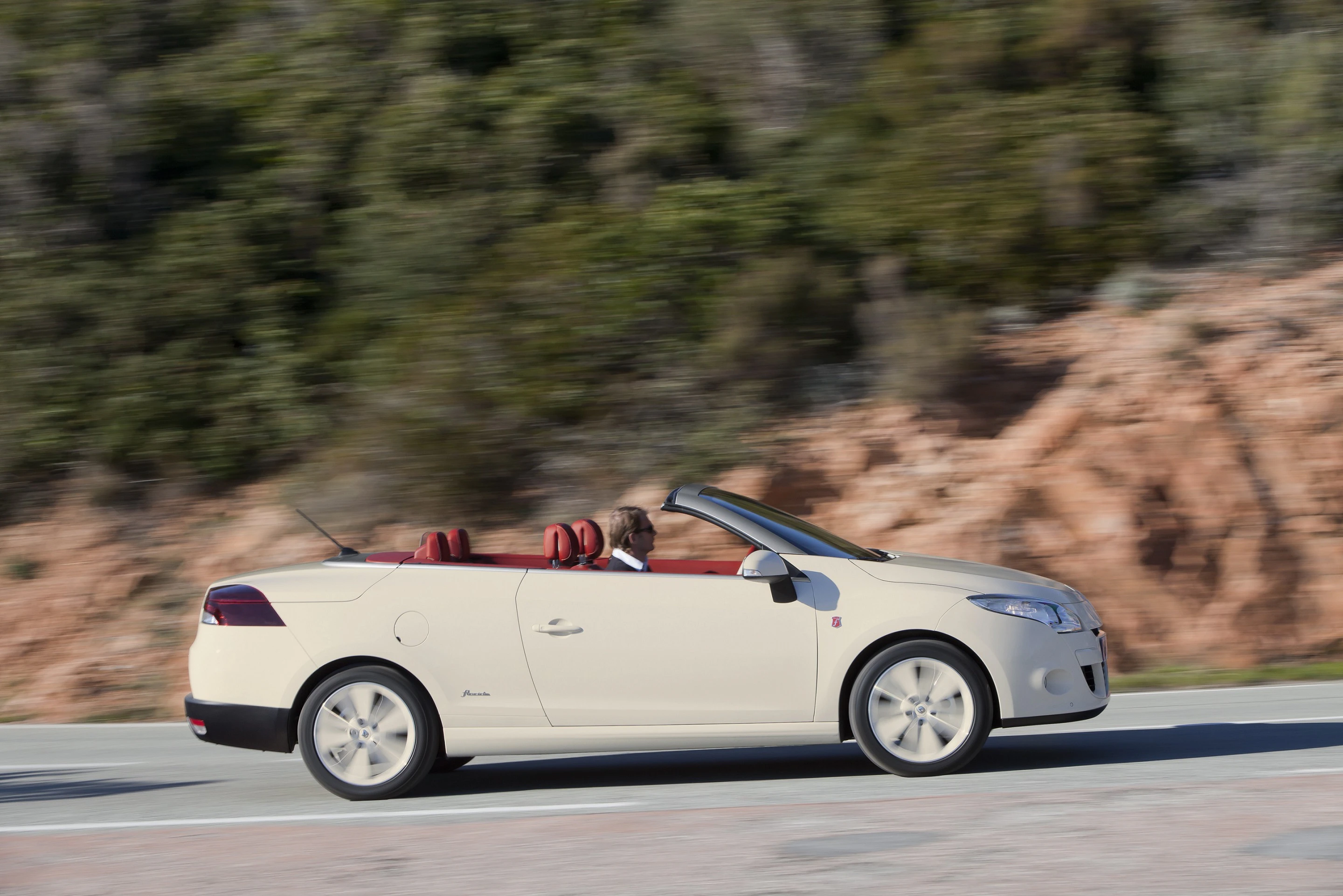 Beiges Cabriolet, fährt auf einer Straße durch Landschaft mit Felsen und Bäumen. Fahrer im Auto, Verdeck offen.