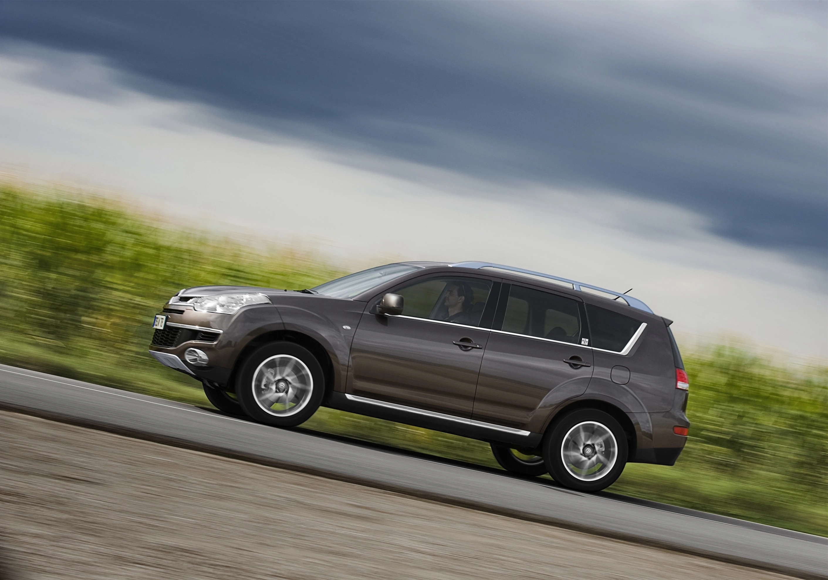 SUV fährt schnell auf einer Landstraße, verschwommener Hintergrund mit Wolken und grüner Vegetation, sportlich-elegantes Design.