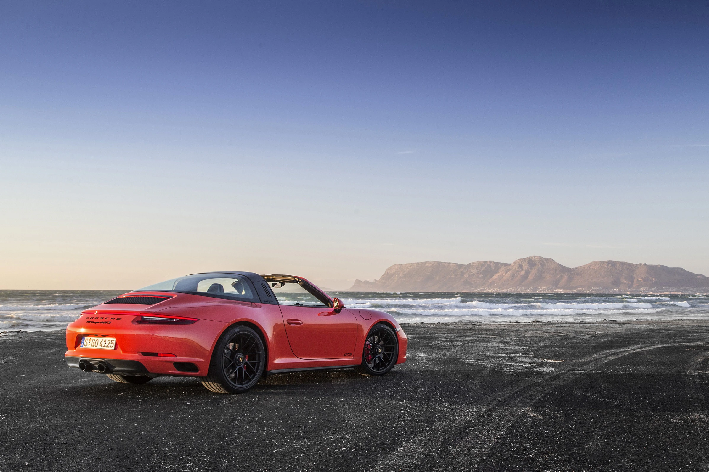 Roter Sportwagen steht am Strand bei Sonnenuntergang, im Hintergrund Berge und Meer.