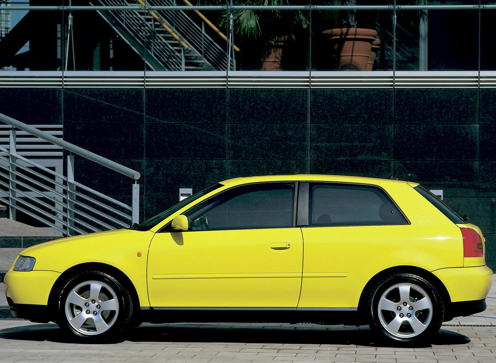 Gelbes Auto parkt vor einem modernen Gebäude mit Glasfassade und Treppen. Seitenansicht zeigt klare Linien und sportliches Design.