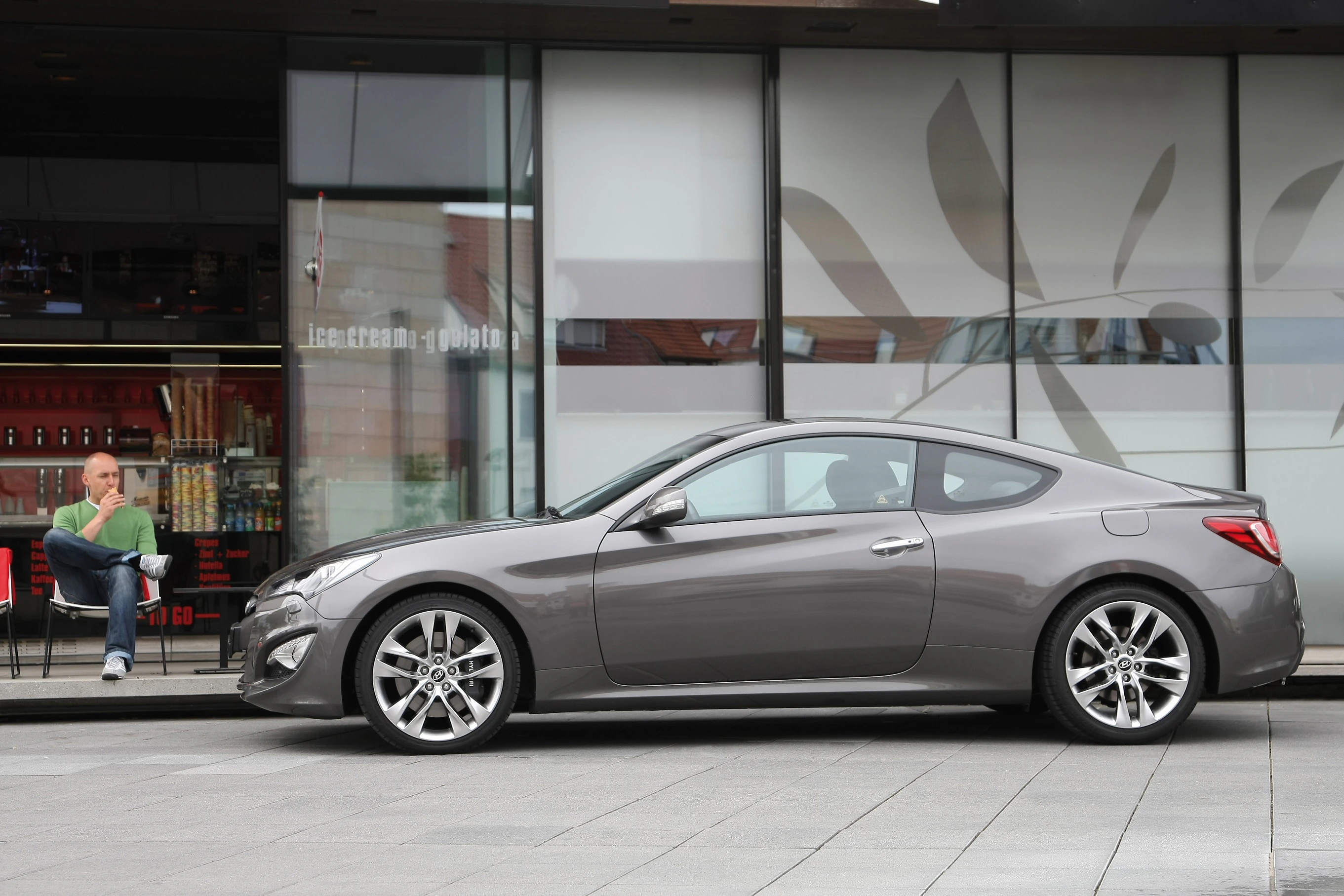 Graues Sportcoupé parkt vor einem modernen Gebäude. Mann sitzt im Hintergrund auf einem Stuhl, neben einem Glastüren-Eingang.