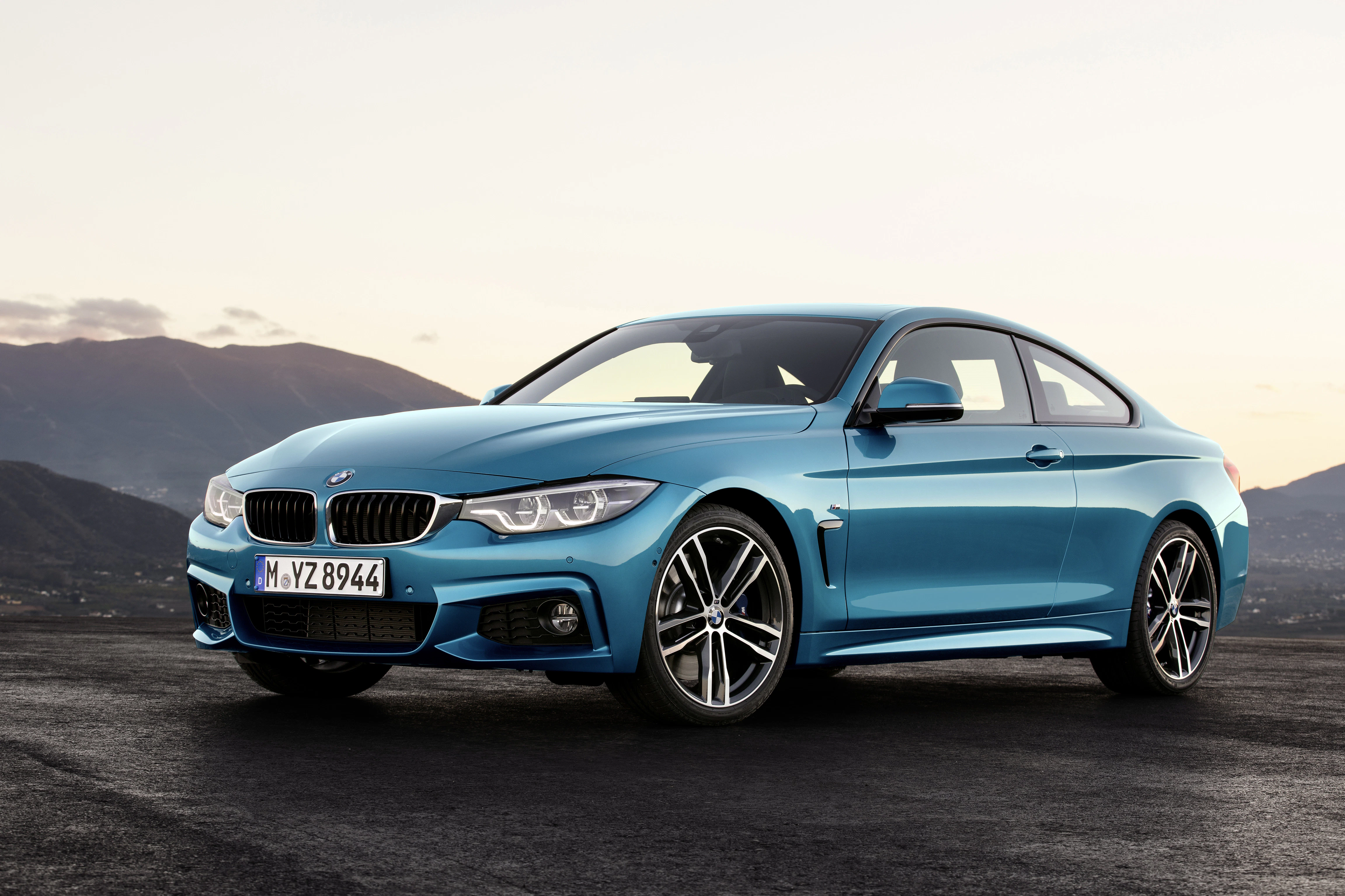 Ein blauer BMW Coupé steht auf einer Straße vor einer bergigen Landschaft bei Sonnenuntergang.