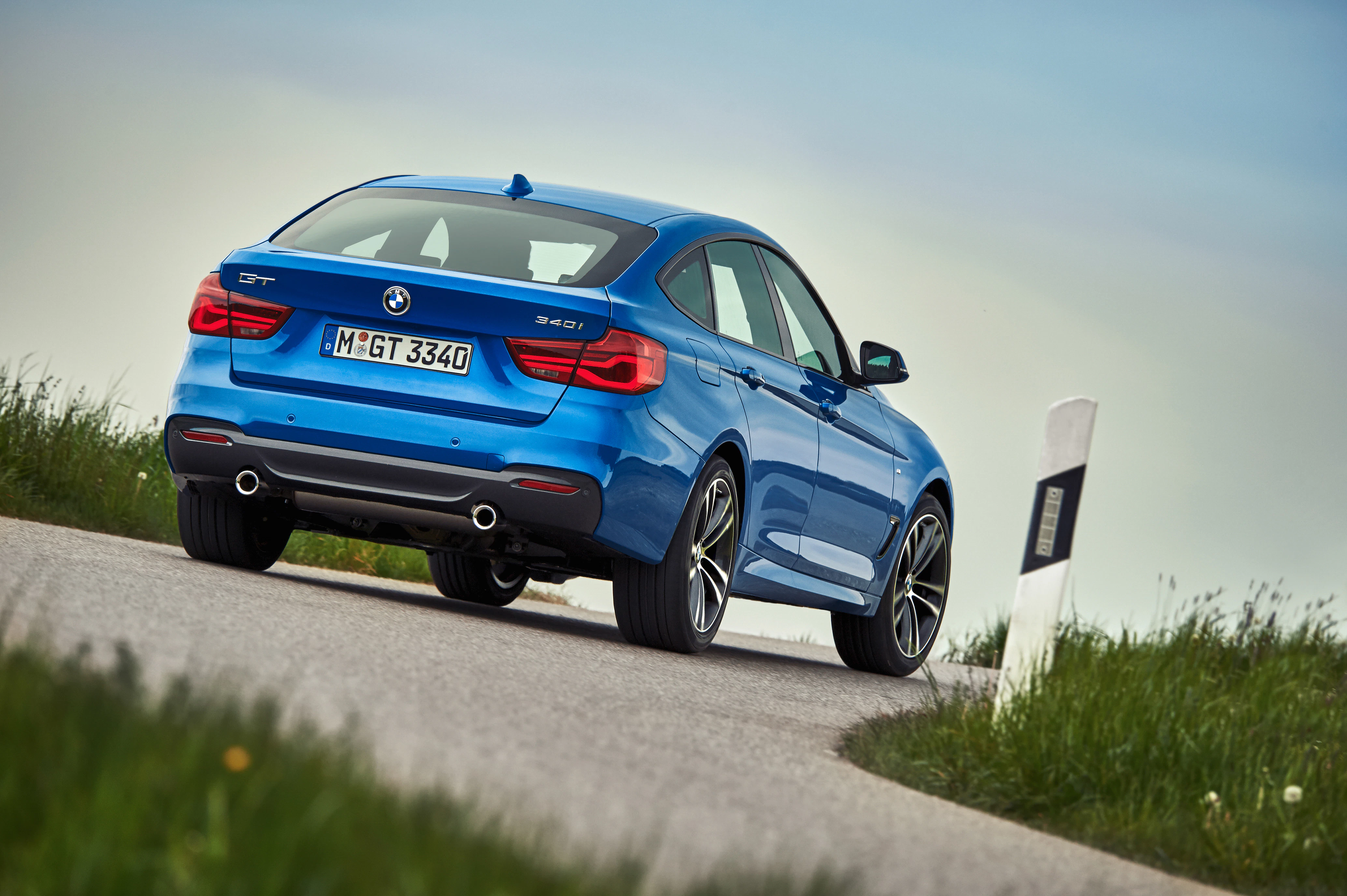 Blauer BMW, Modell 3er GT, fährt auf einer ländlichen Straße mit grünen Wiesen an den Seiten unter blauem Himmel.