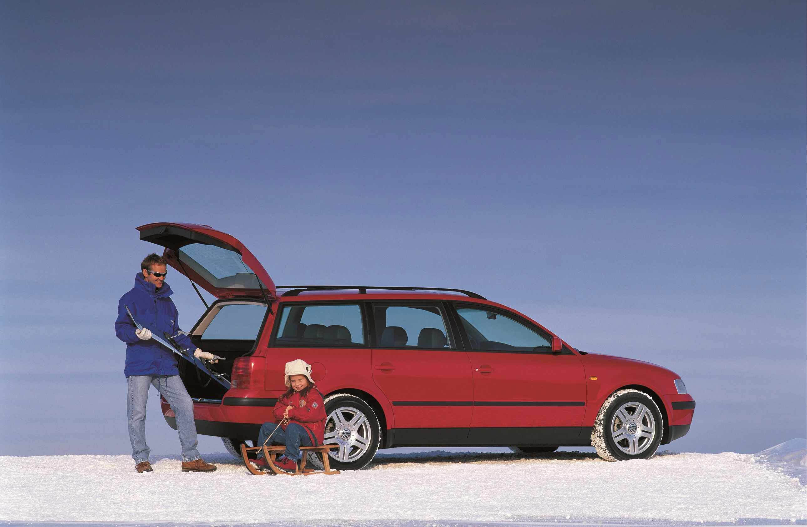 Ein Mann steht neben einem roten Kombi auf Schnee, während ein Kind auf einem Schlitten sitzt. Der Kofferraum des Autos ist geöffnet.
