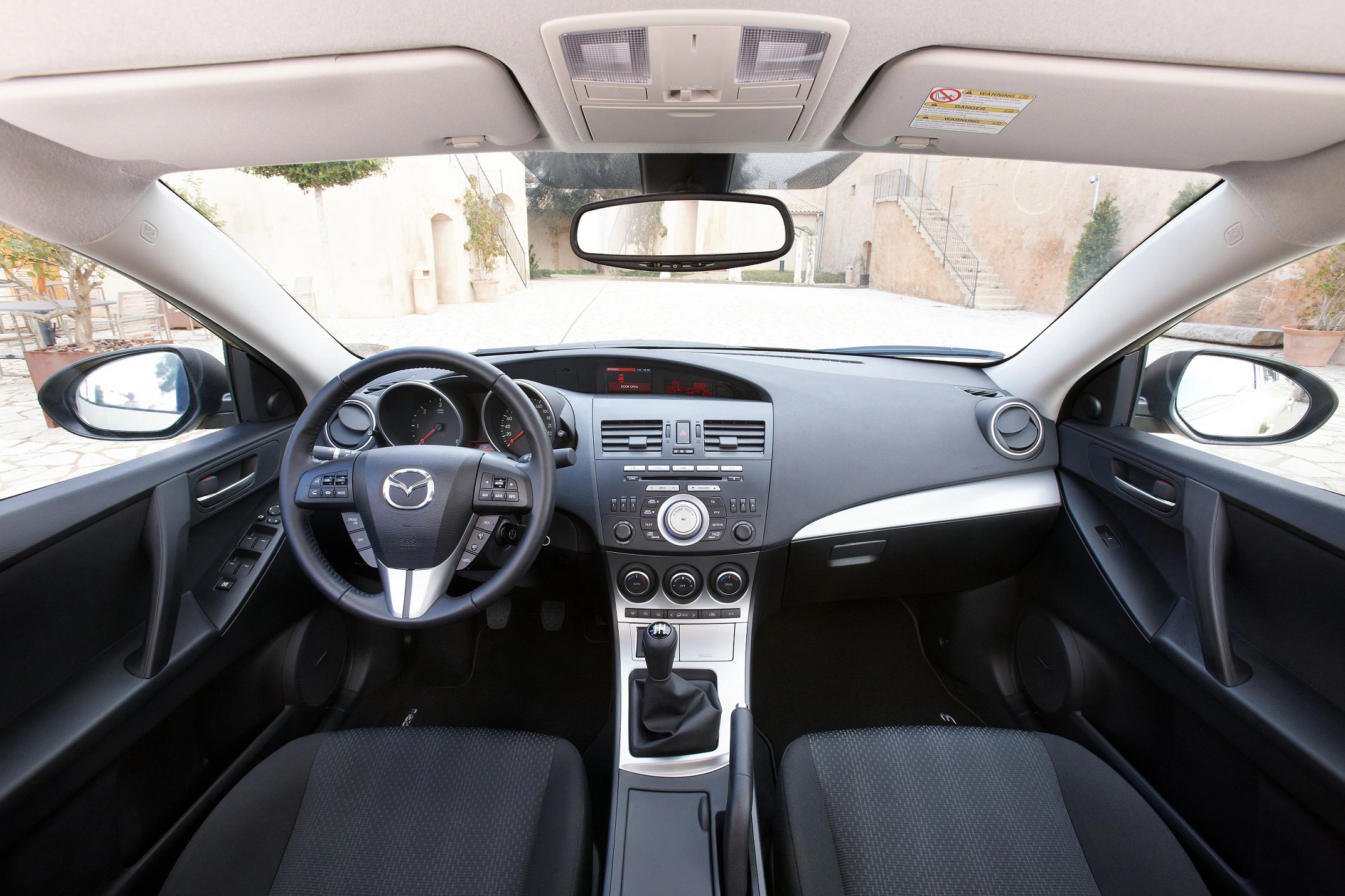 Innenraum eines Mazda-Fahrzeugs mit schwarzer Ausstattung, zentralem Armaturenbrett, Lenkrad links und Schaltknüppel in der Mitte.