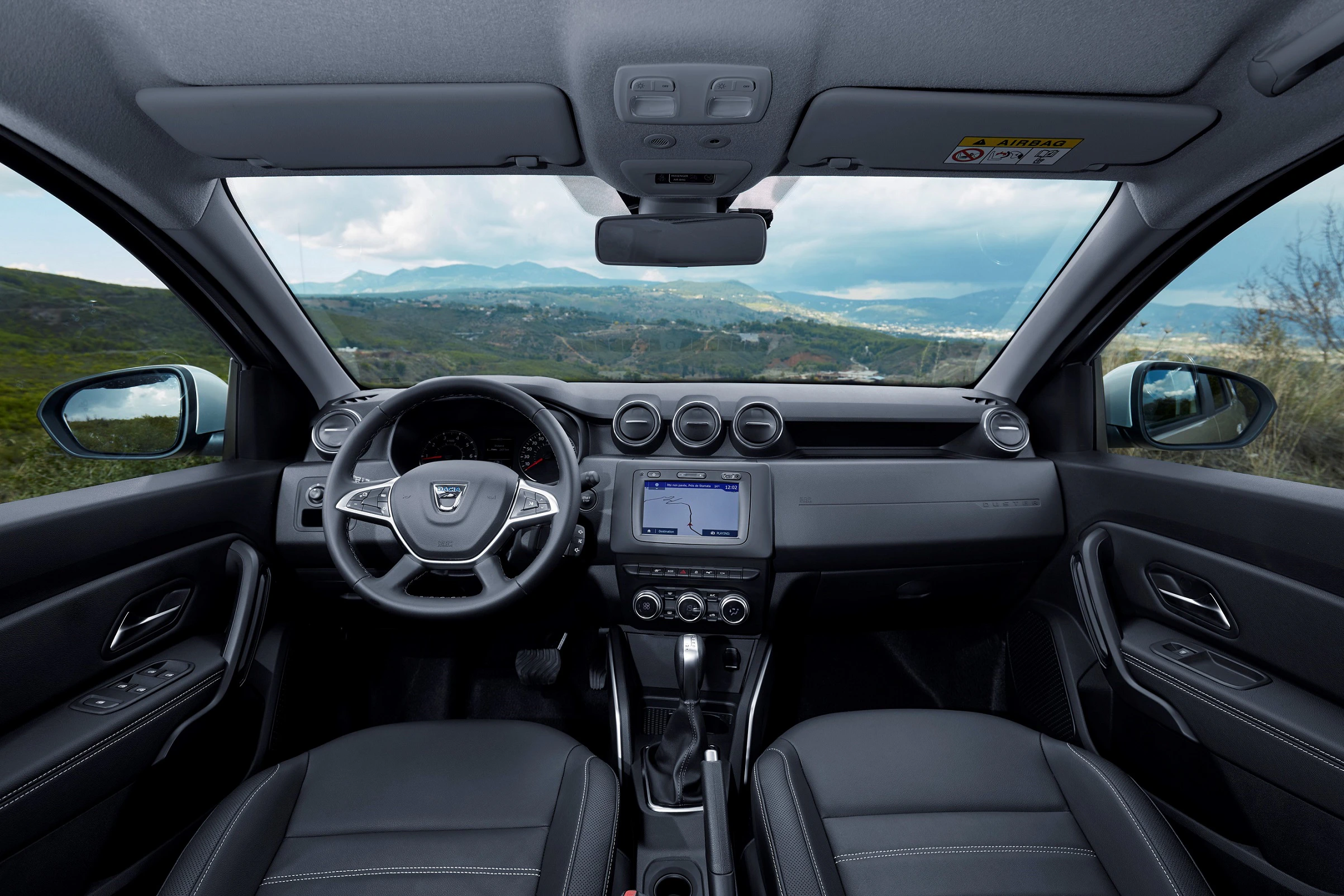 Innenraum eines Autos mit Blick auf ein modernes Armaturenbrett, ein Lenkrad und ein Touchscreen-Display. Draußen ist eine Landschaft sichtbar.