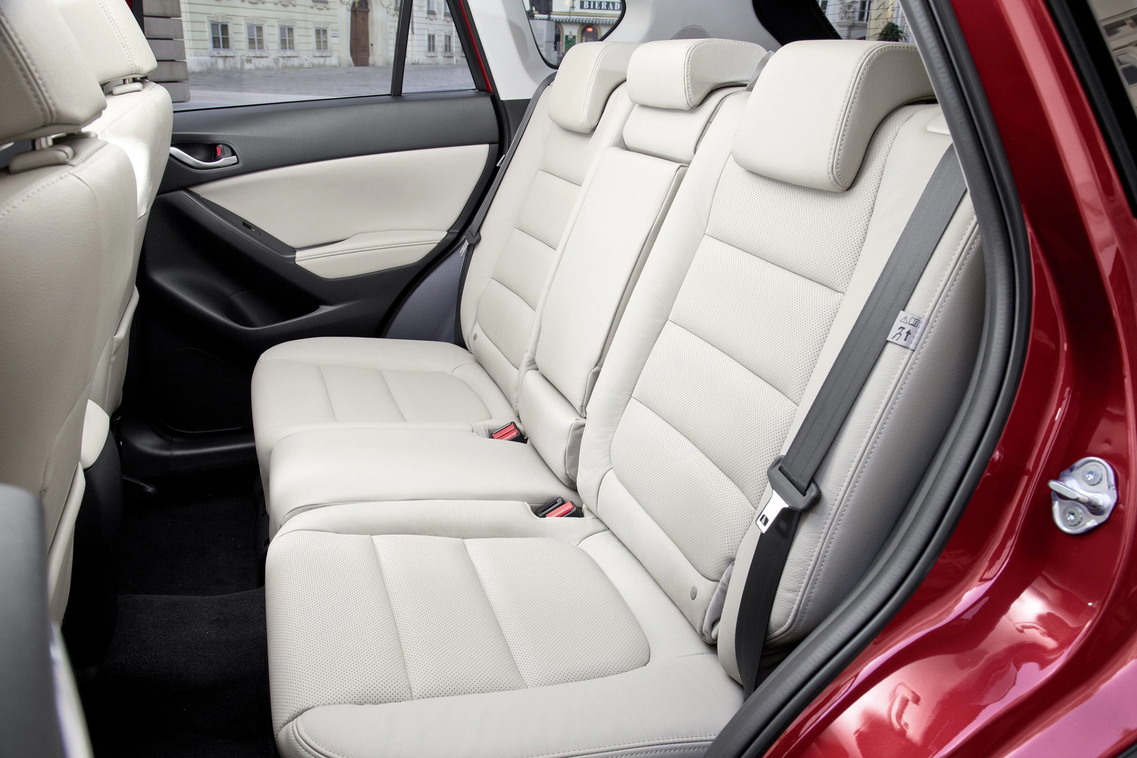 Rückbank eines Autos mit hellen Ledersitzen, drei Sitzplätzen mit Gurten, in einem modernen, roten Fahrzeuginterieur.