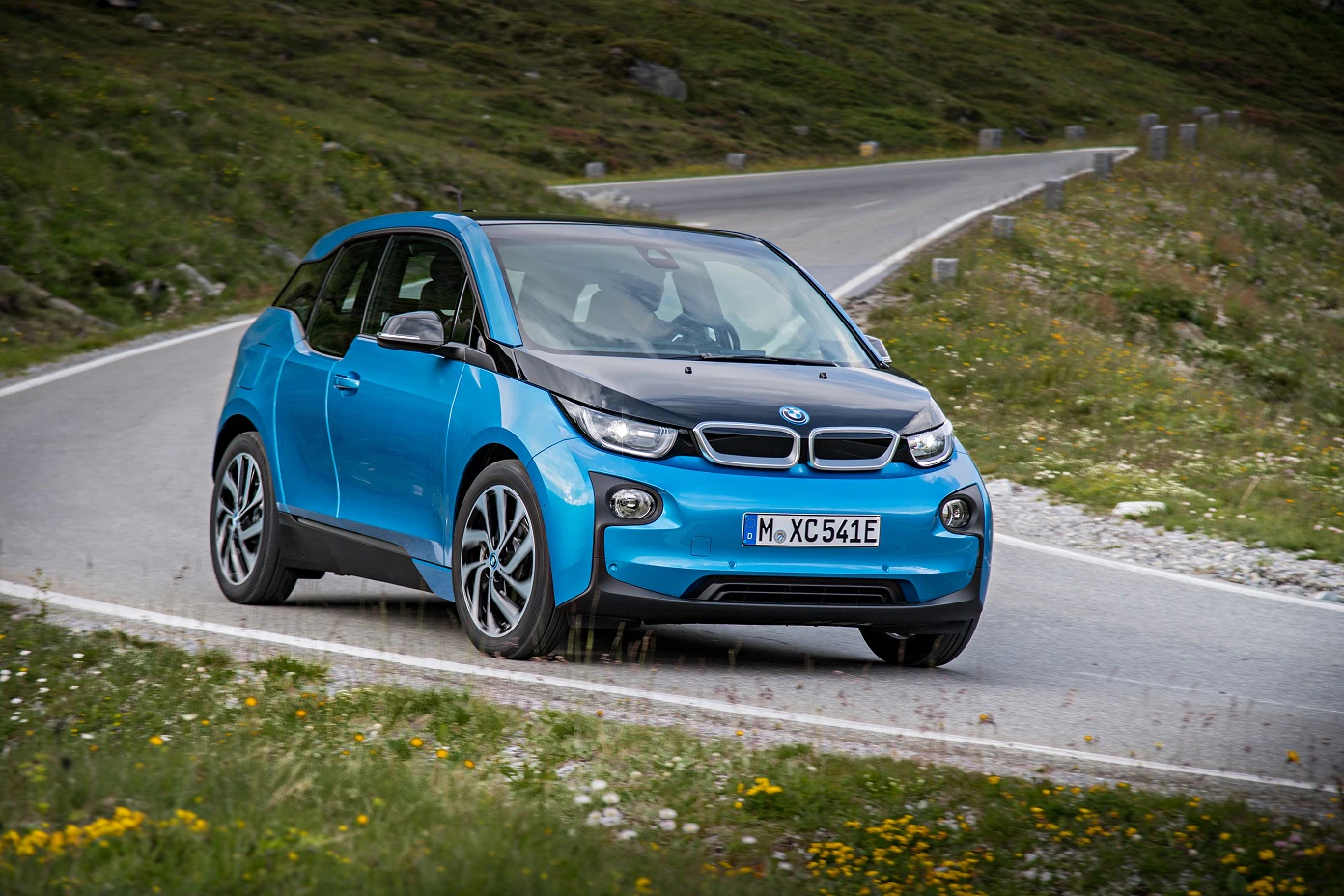 Blauer BMW i3 fährt auf einer kurvigen Landstraße durch eine grüne, bergige Landschaft.