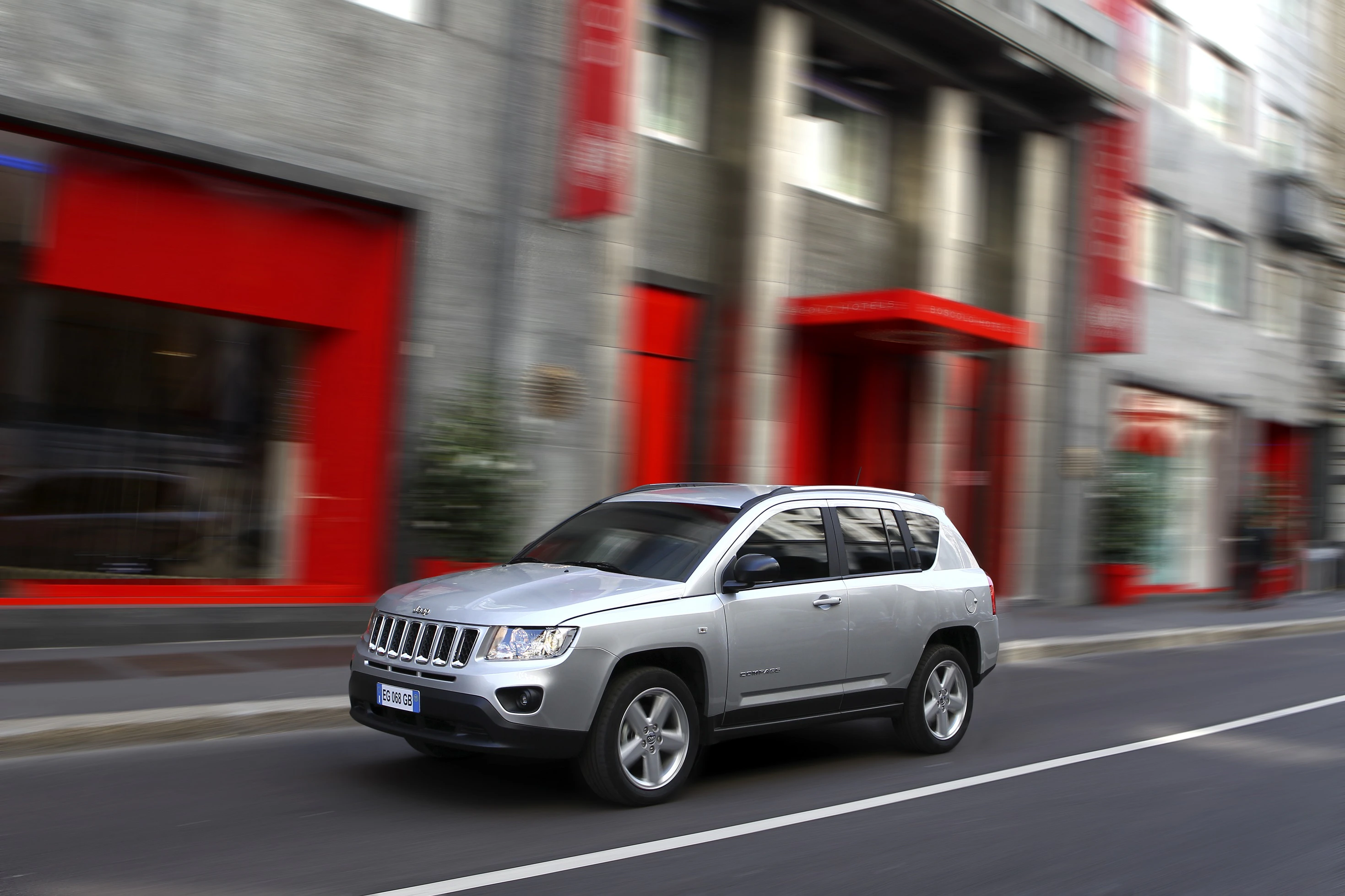 Silberner SUV fährt auf städtischer Straße mit unscharfen roten und grauen Gebäuden im Hintergrund.