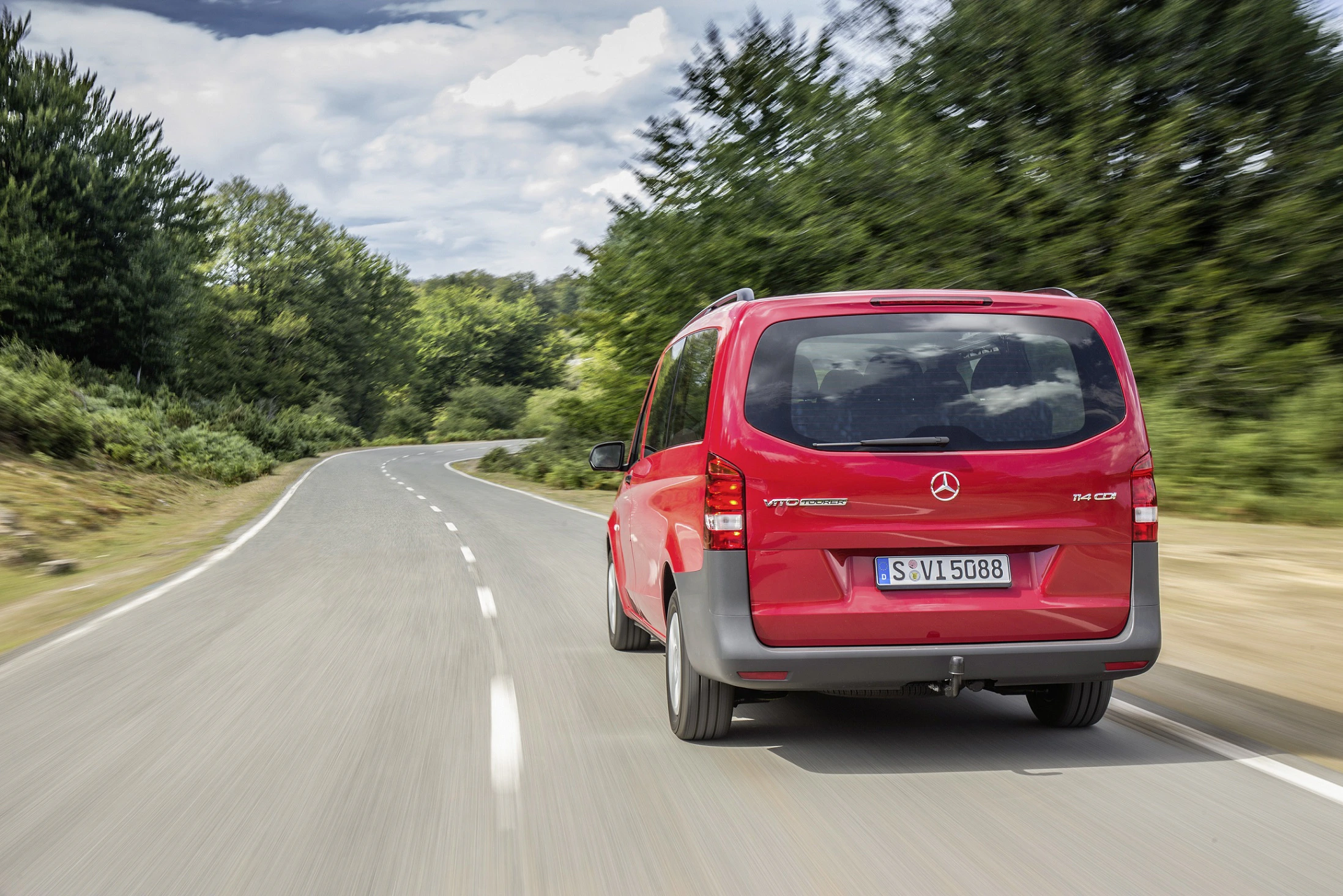 Roter Mercedes-Benz Van fährt auf einer kurvigen Landstraße, umgeben von Bäumen und bewölktem Himmel.