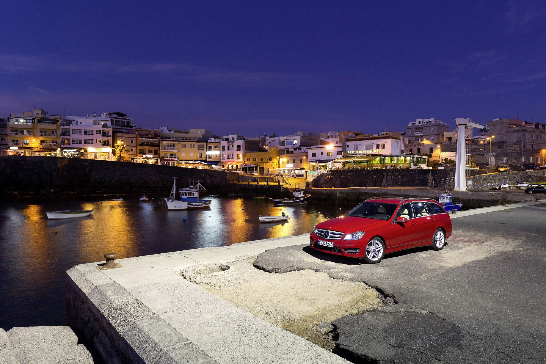 Roter Kombi parkt am Hafen bei Nacht, beleuchtete Gebäude im Hintergrund und Boote auf dem Wasser im Vordergrund.