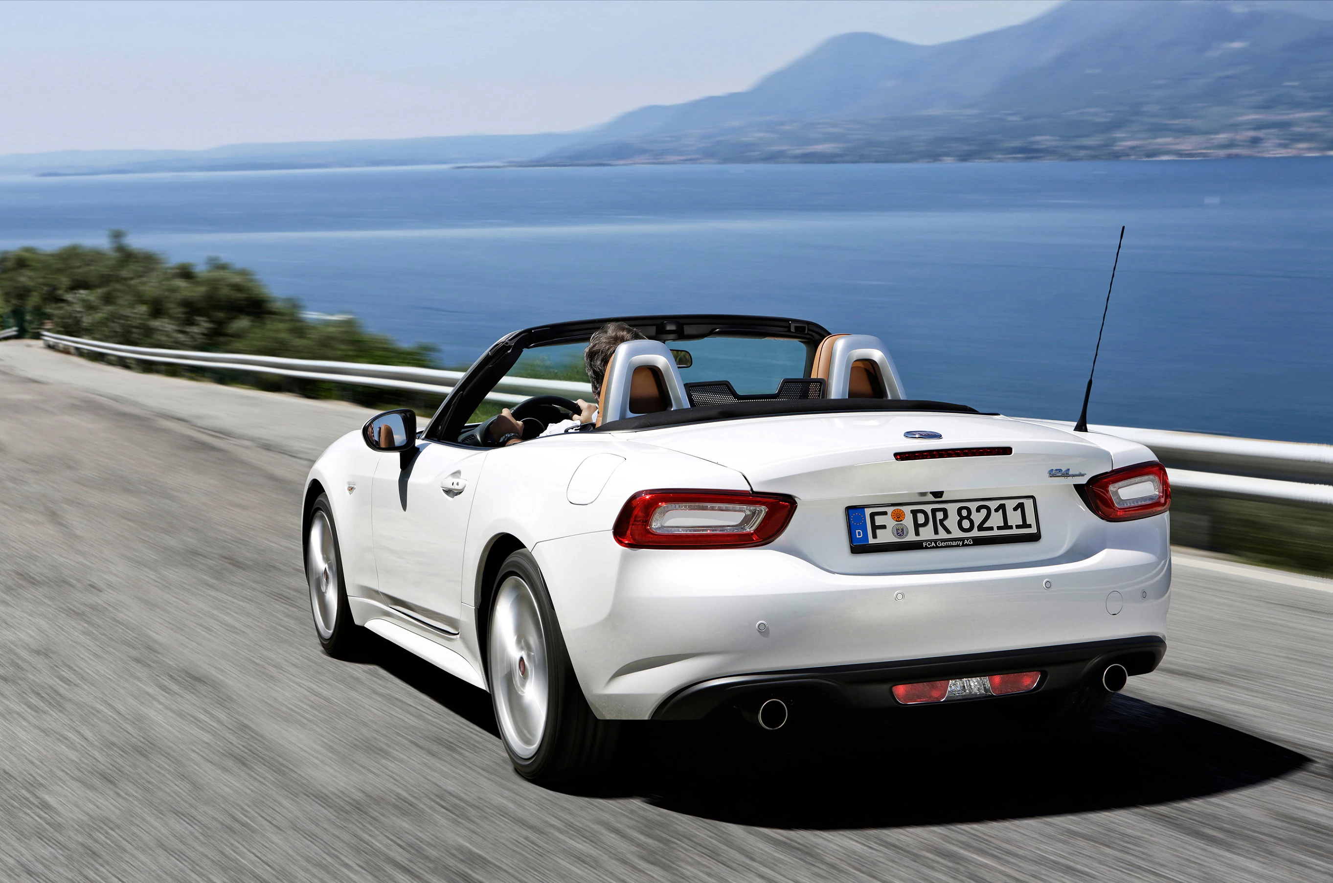 Weißes Cabrio fährt auf Küstenstraße entlang des Meeres, mit bewaldeten Hügeln im Hintergrund bei sonnigem Wetter.