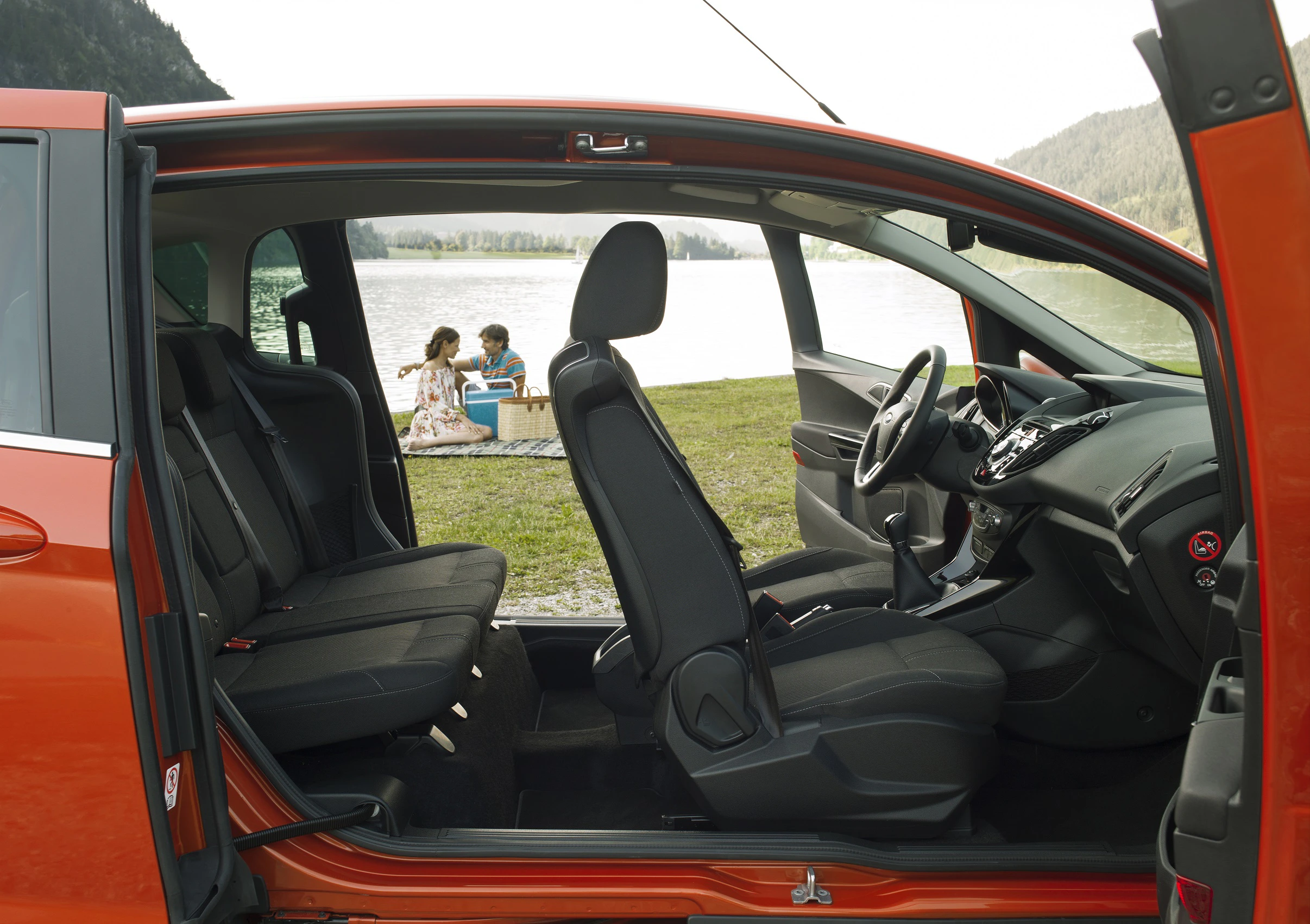 Innenansicht eines roten Autos mit geöffneter Tür. Im Hintergrund sitzen zwei Personen am Ufer eines Sees.