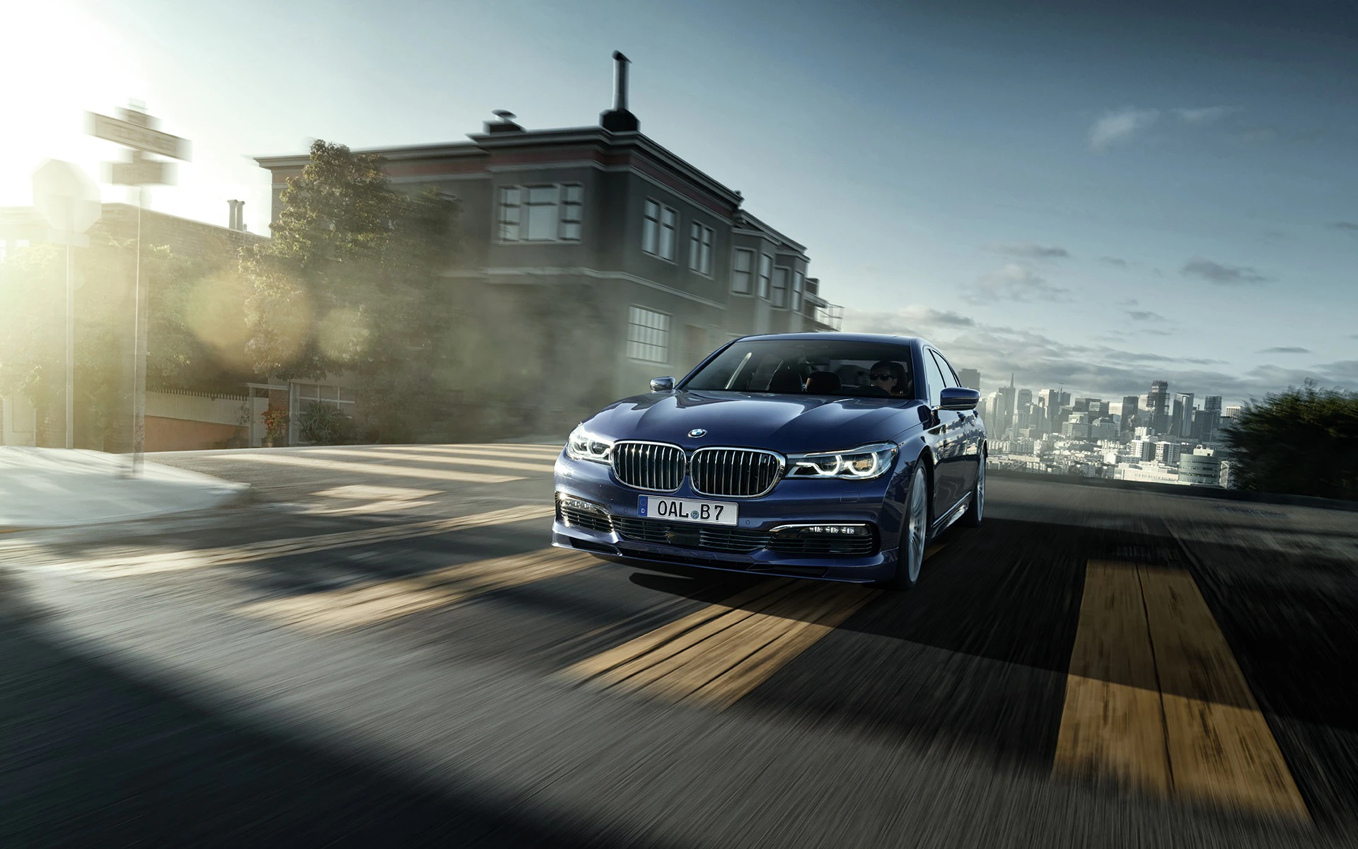 Blauer BMW fährt in städtischer Umgebung, im Vordergrund eine gelbe Straßenschwelle, im Hintergrund Gebäude und Skyline bei bewölktem Himmel.