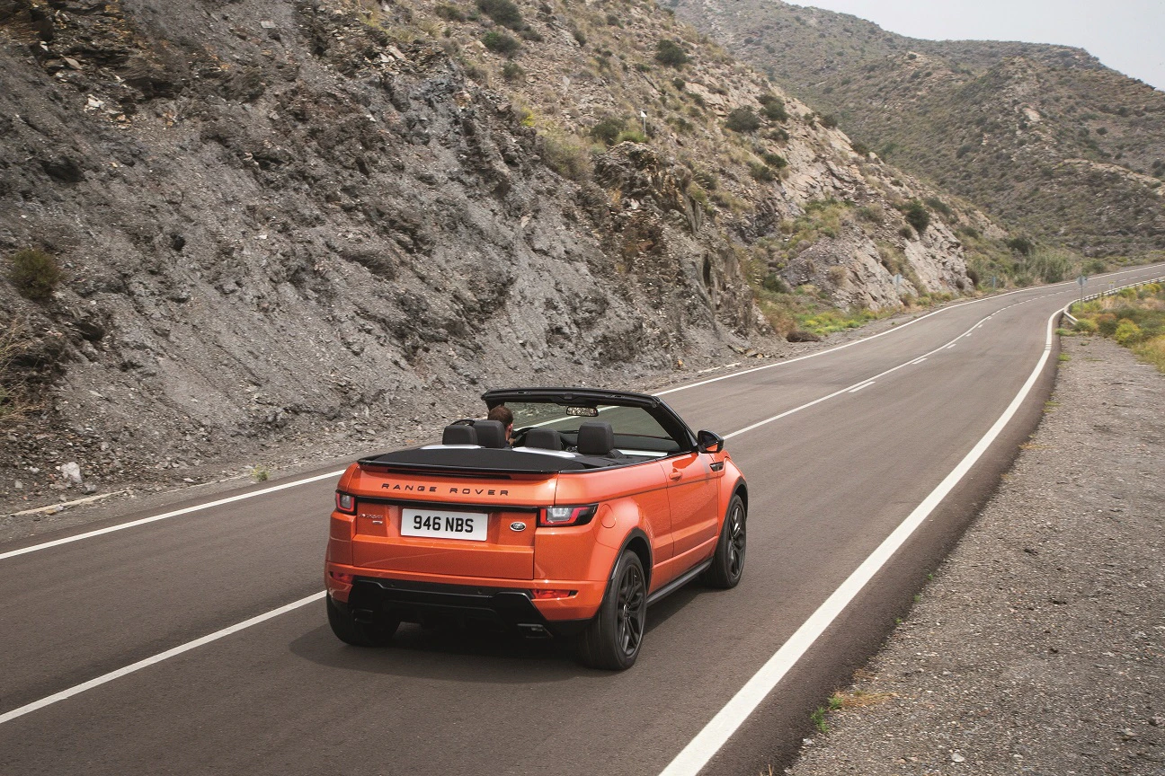 Ein orangefarbener Land Rover Cabrio fährt auf einer kurvenreichen Bergstraße an einer felsigen Landschaft entlang.