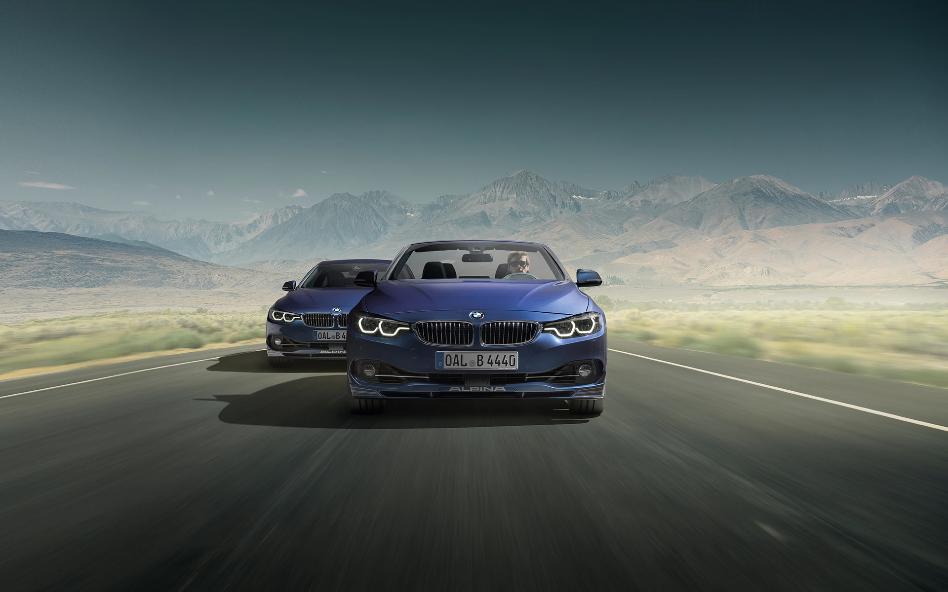 Zwei blaue BMWs fahren auf einer offenen Straße durch eine malerische Berglandschaft unter klarem Himmel.