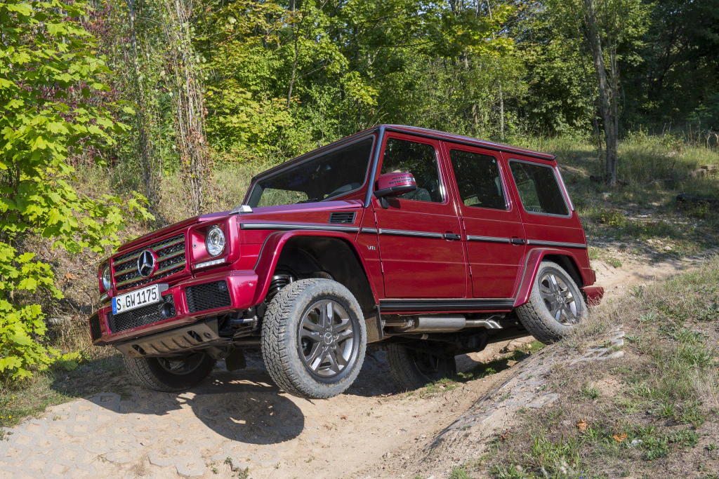 Roter Mercedes-Geländewagen fährt Offroad auf unebenem, sandigem Gelände mit grüner Vegetation im Hintergrund.