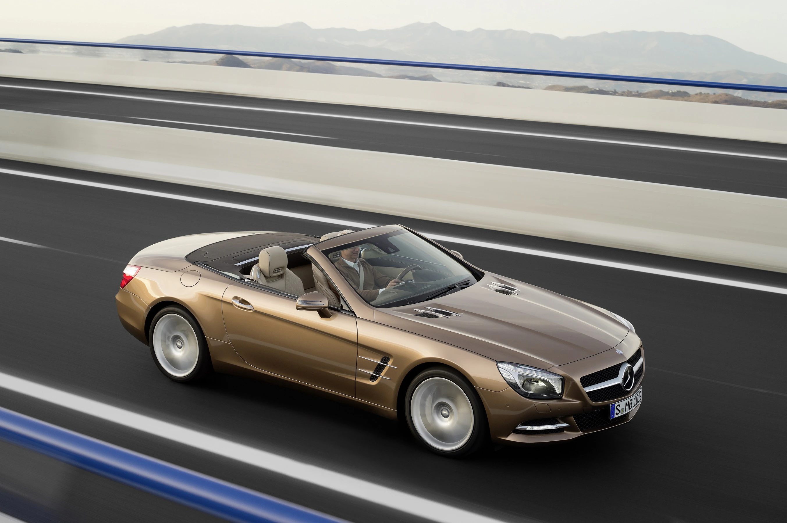 Brauner Mercedes-Benz Cabriolet fährt auf einer Autobahnbrücke bei klarem Wetter, im Hintergrund sind Berge zu sehen.