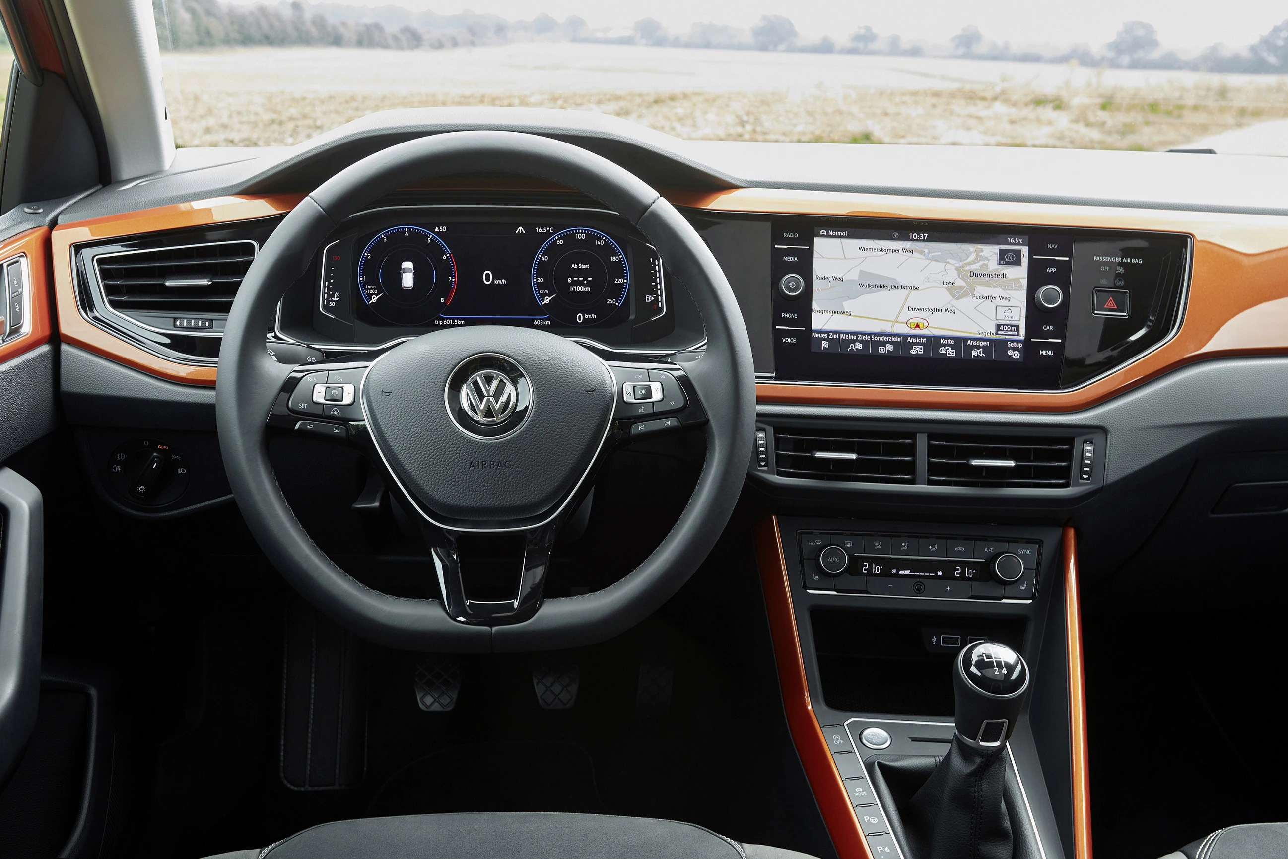 Innenraum eines modernen Autos mit Lenkrad, Digitaltacho, Navigationsbildschirm und manuellem Schalthebel, orange Akzente.