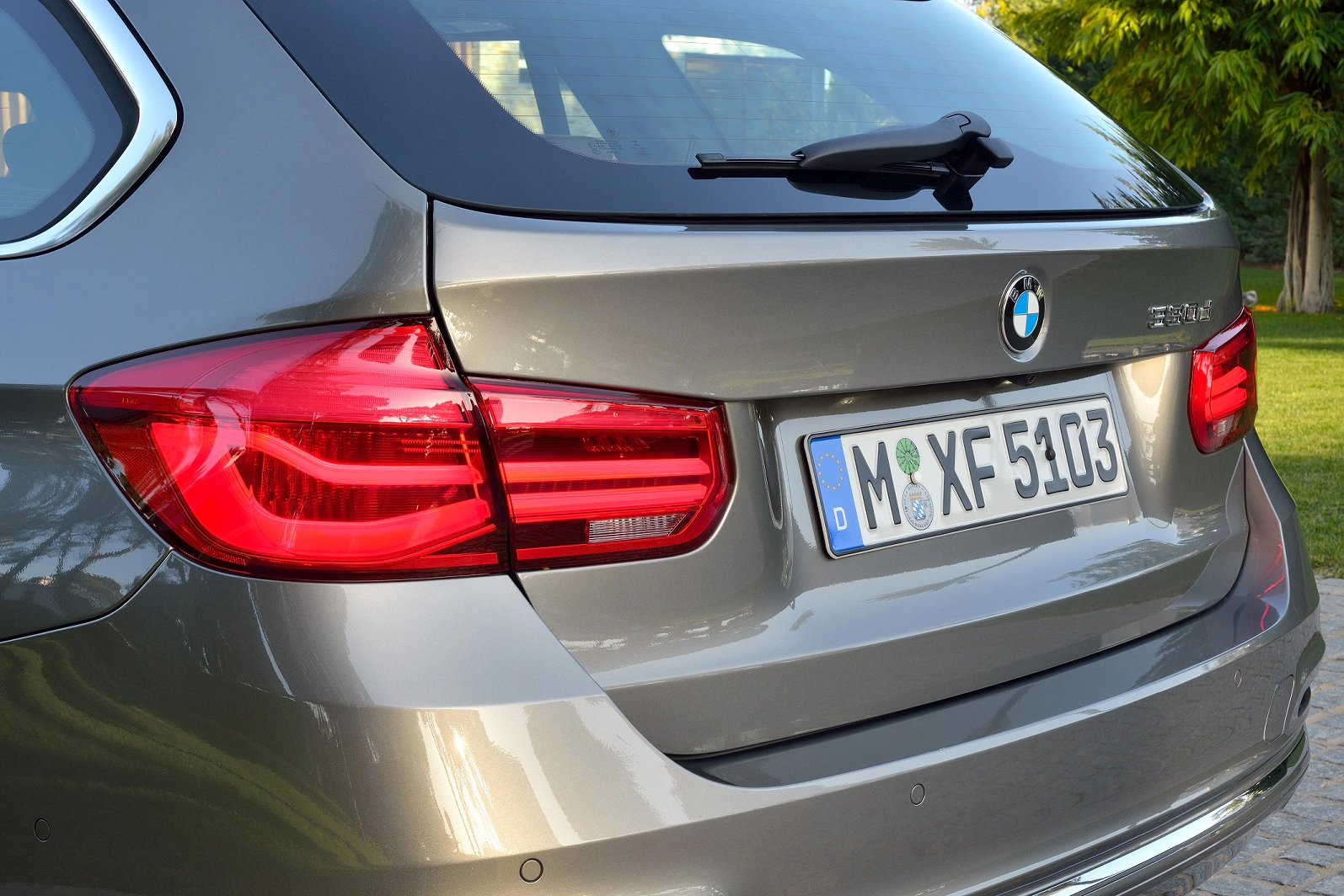 Rückansicht eines grauen BMW 216d mit deutschem Kennzeichen, geparktem Auto im Freien vor einer grünen Hecke.