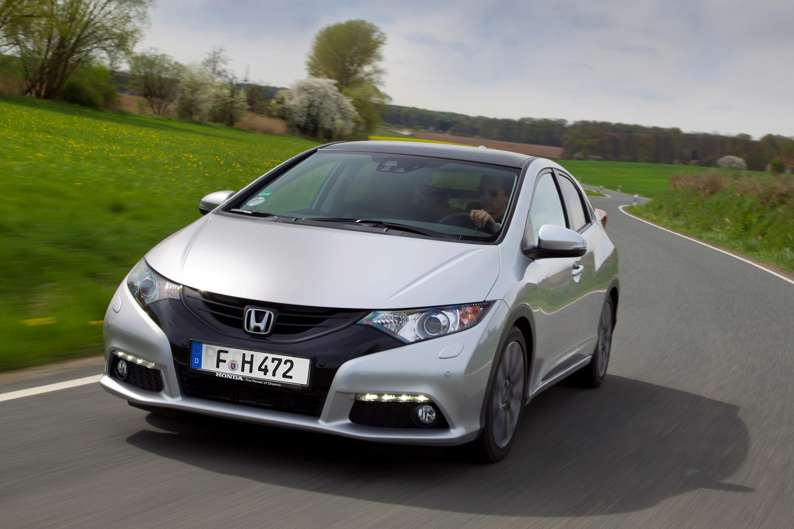 Silberner Honda Civic fährt auf einer ländlichen Straße mit grünen Wiesen und Bäumen im Hintergrund.