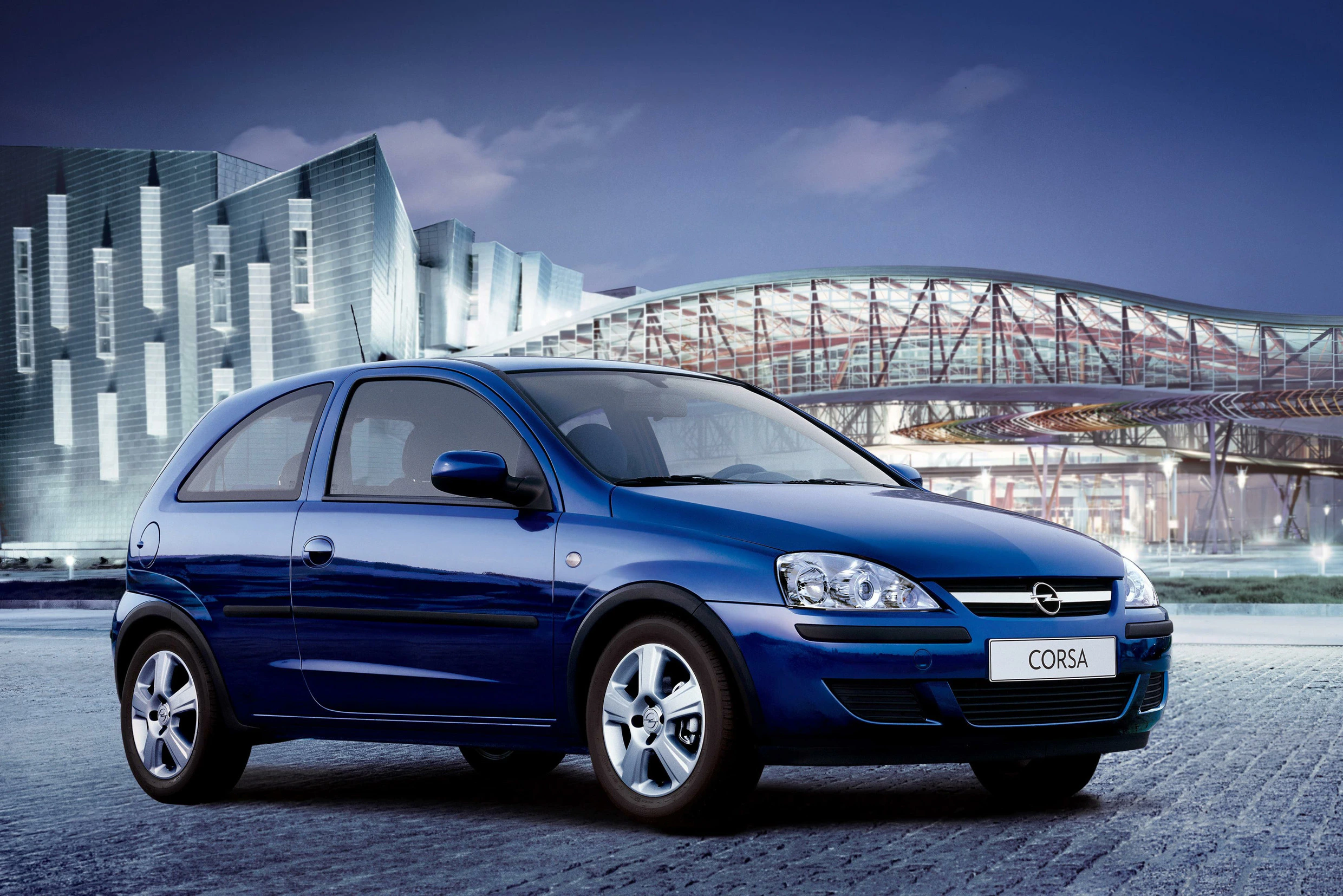 Blauer Opel Corsa steht auf gepflastertem Platz vor moderner Architektur bei Dämmerung.