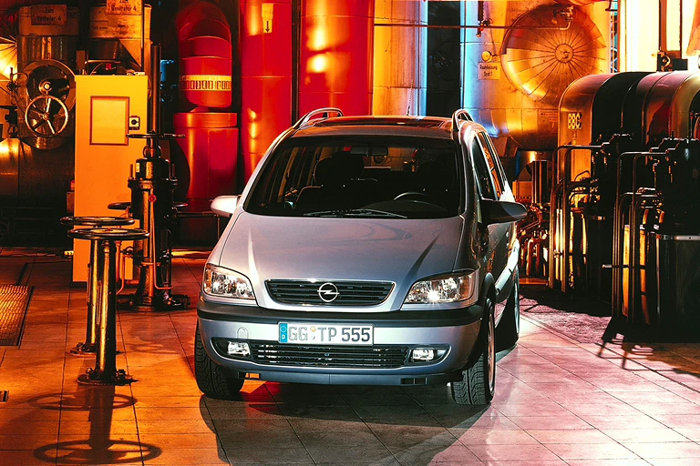 Silberner Opel Zafira steht in einer bunt beleuchteten, industriellen Umgebung mit Maschinen und Rohren im Hintergrund.