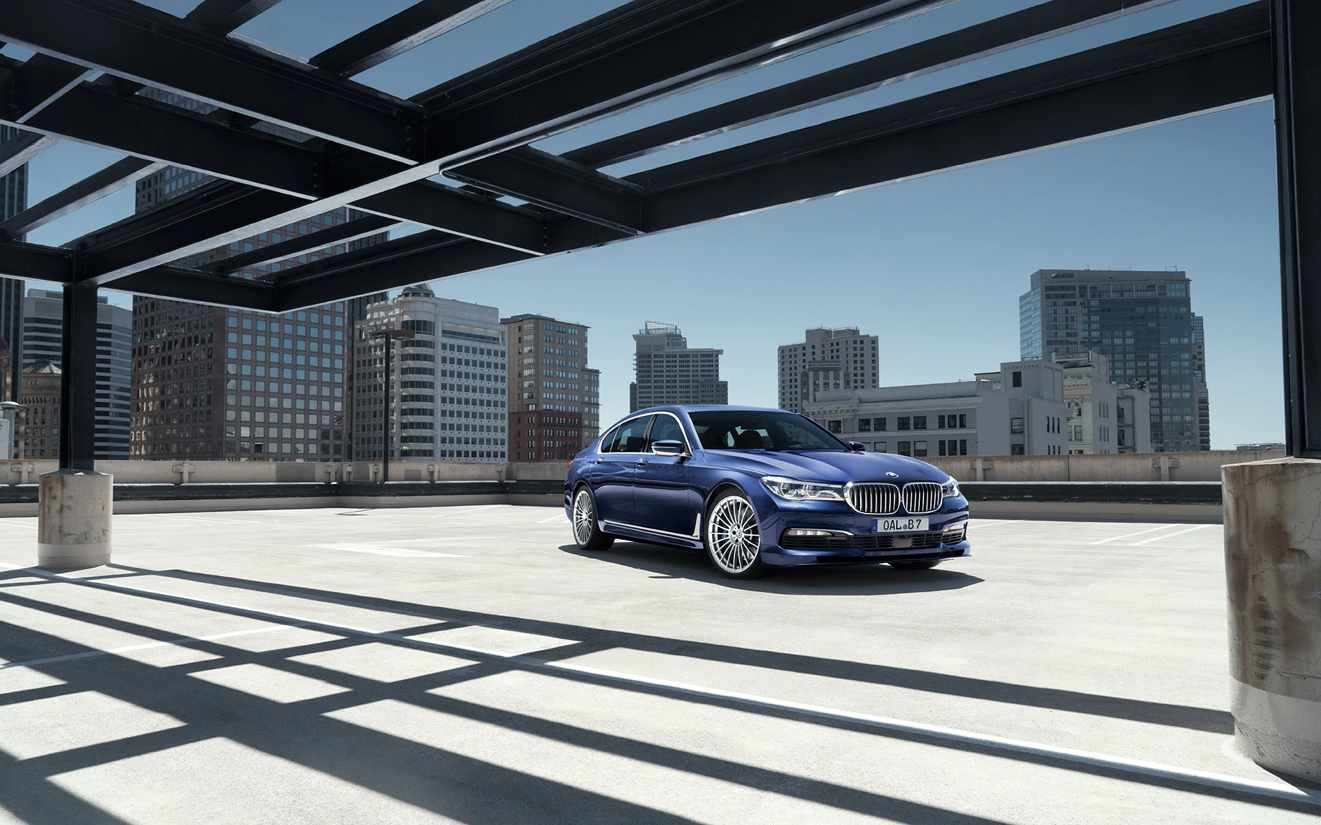 Blauer BMW 7er steht auf einem leeren Parkdeck in einer Stadt, umgeben von Hochhäusern und klarem Himmel.