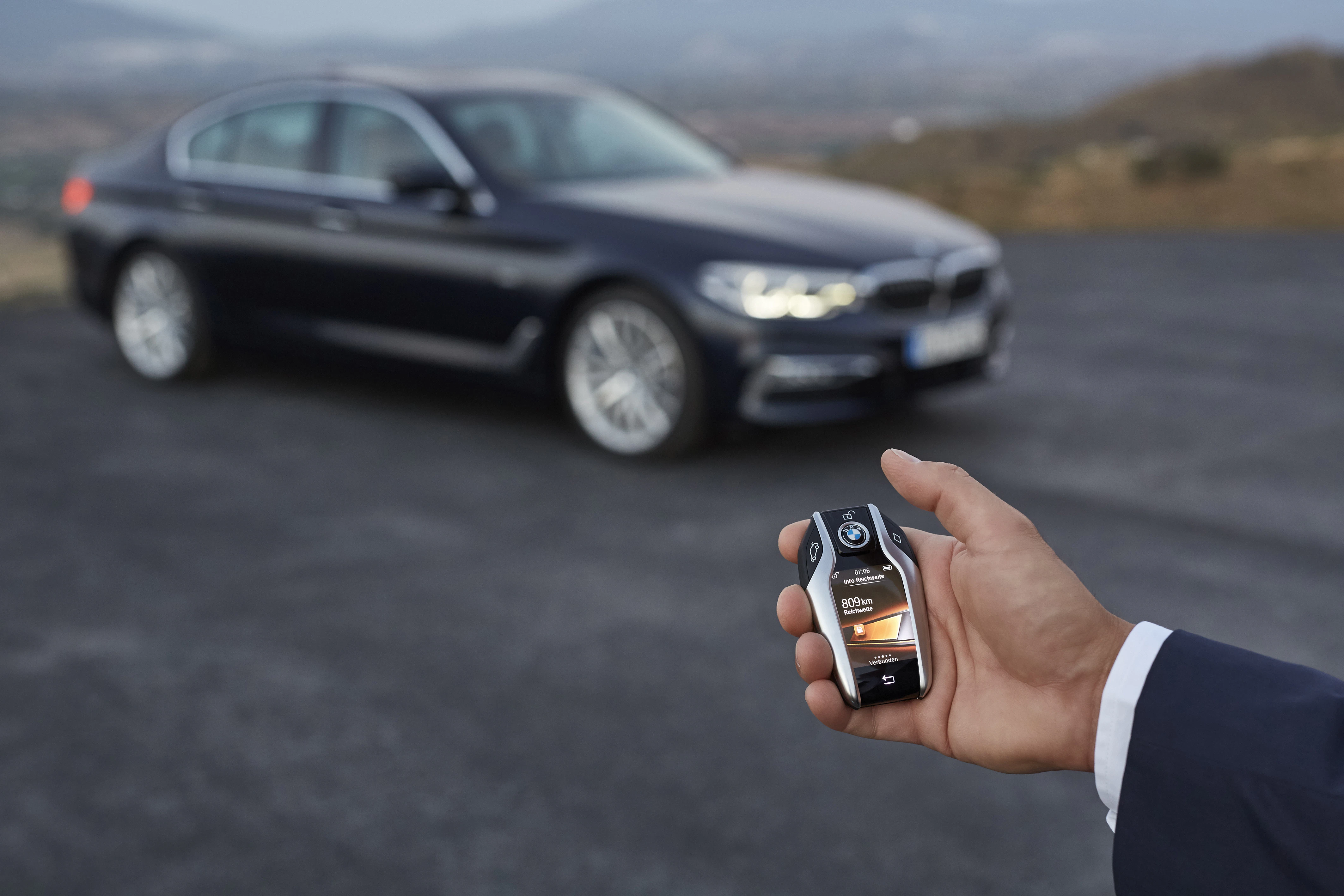 Eine Hand hält einen Autoschlüssel mit Display, im Hintergrund steht ein geparktes schwarzes Auto auf einem Asphaltplatz.