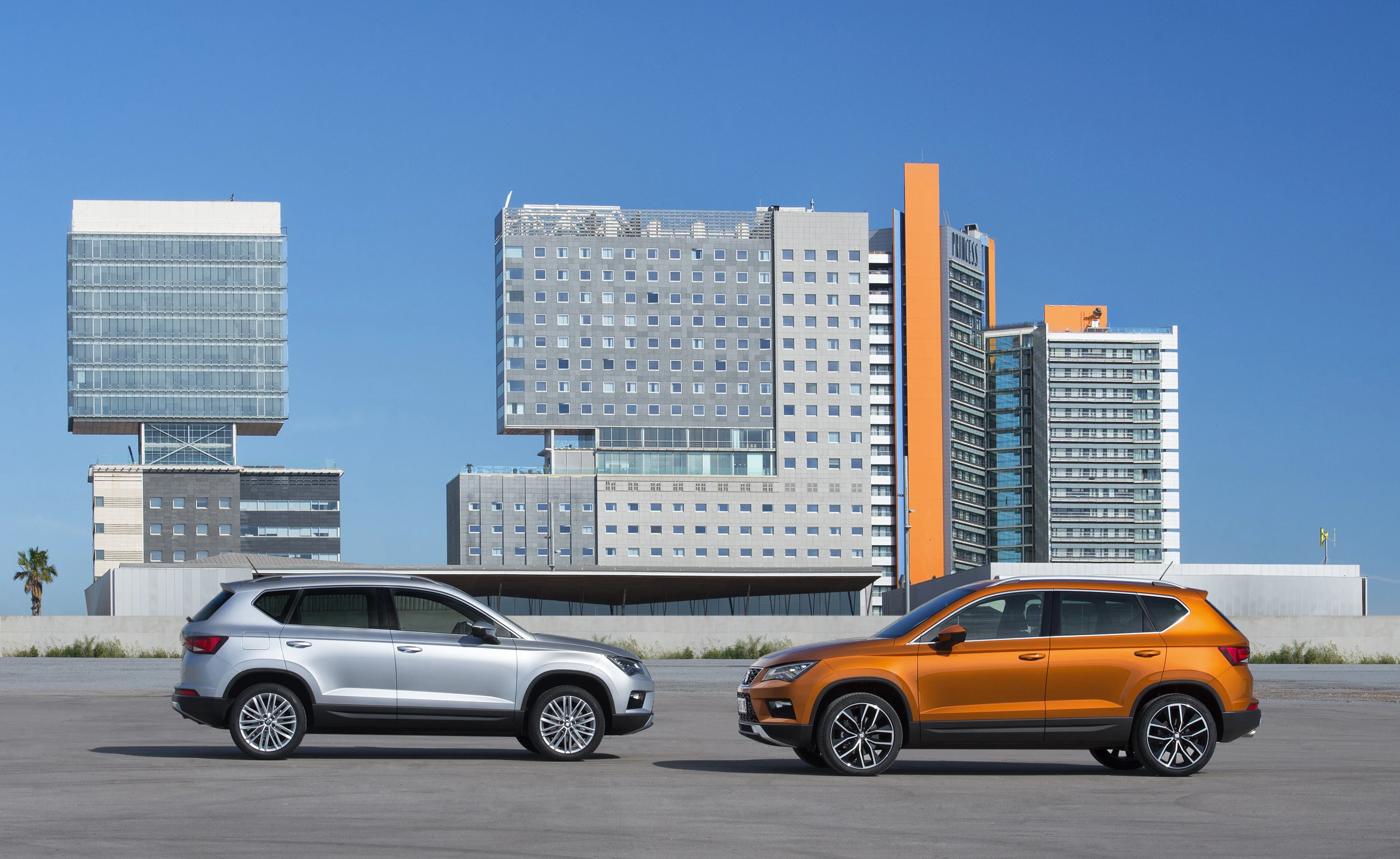 Zwei SUVs, ein silberner und ein orangefarbener, parken nebeneinander vor modernen, hohen Gebäuden bei klarem, blauem Himmel.