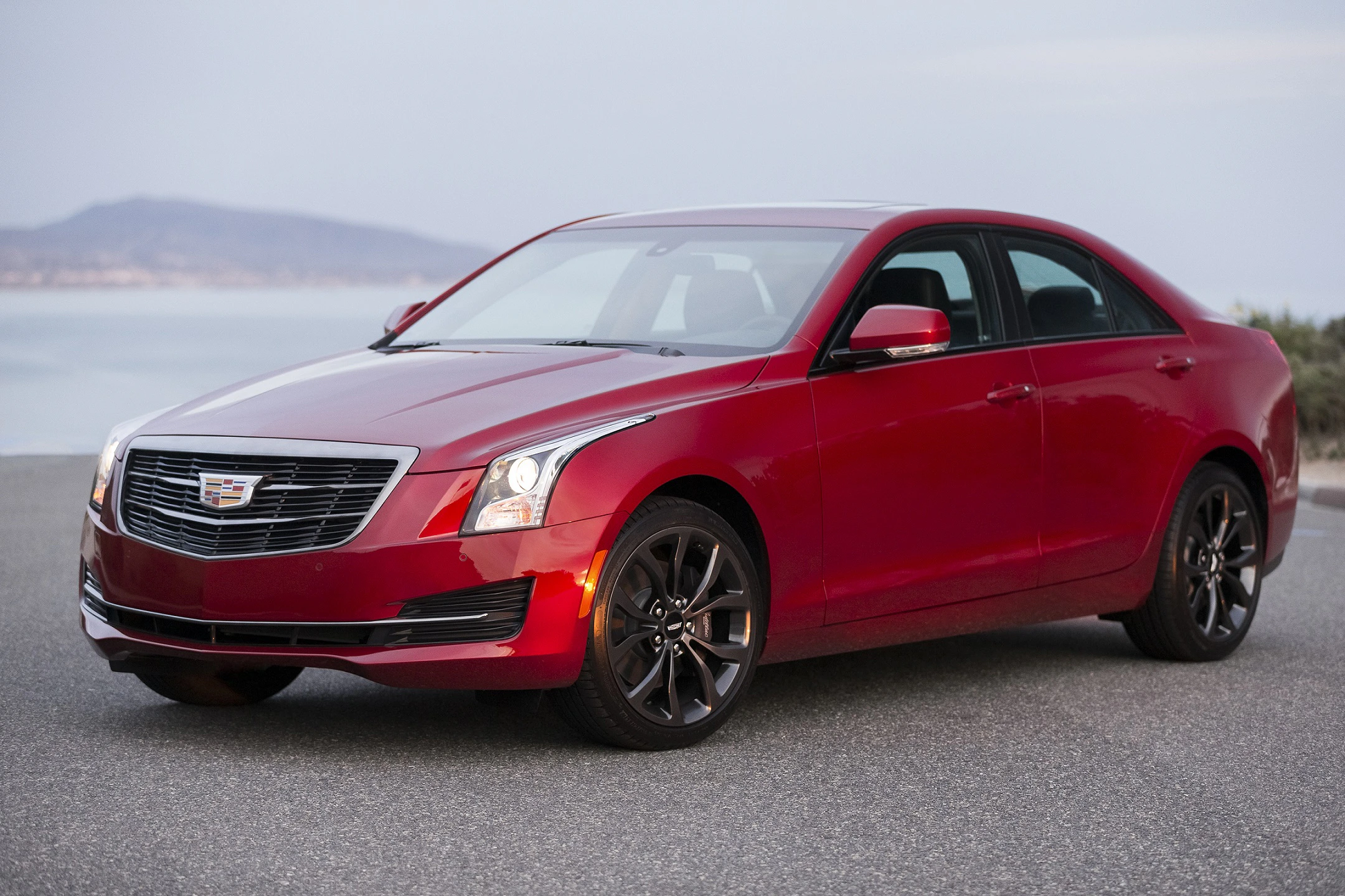 Roter Cadillac ATS Limousine auf einer Küstenstraße bei Sonnenschein; Hintergrund zeigt das Meer und einen Hügel.