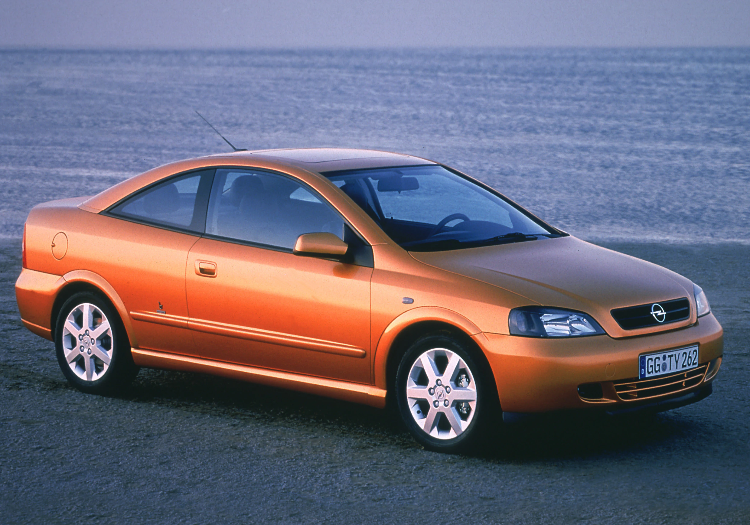 Orangenfarbener Opel Coupé, geparkt am Wasser, seitliche Ansicht, bei bewölktem Himmel. Räder gerade ausgerichtet.