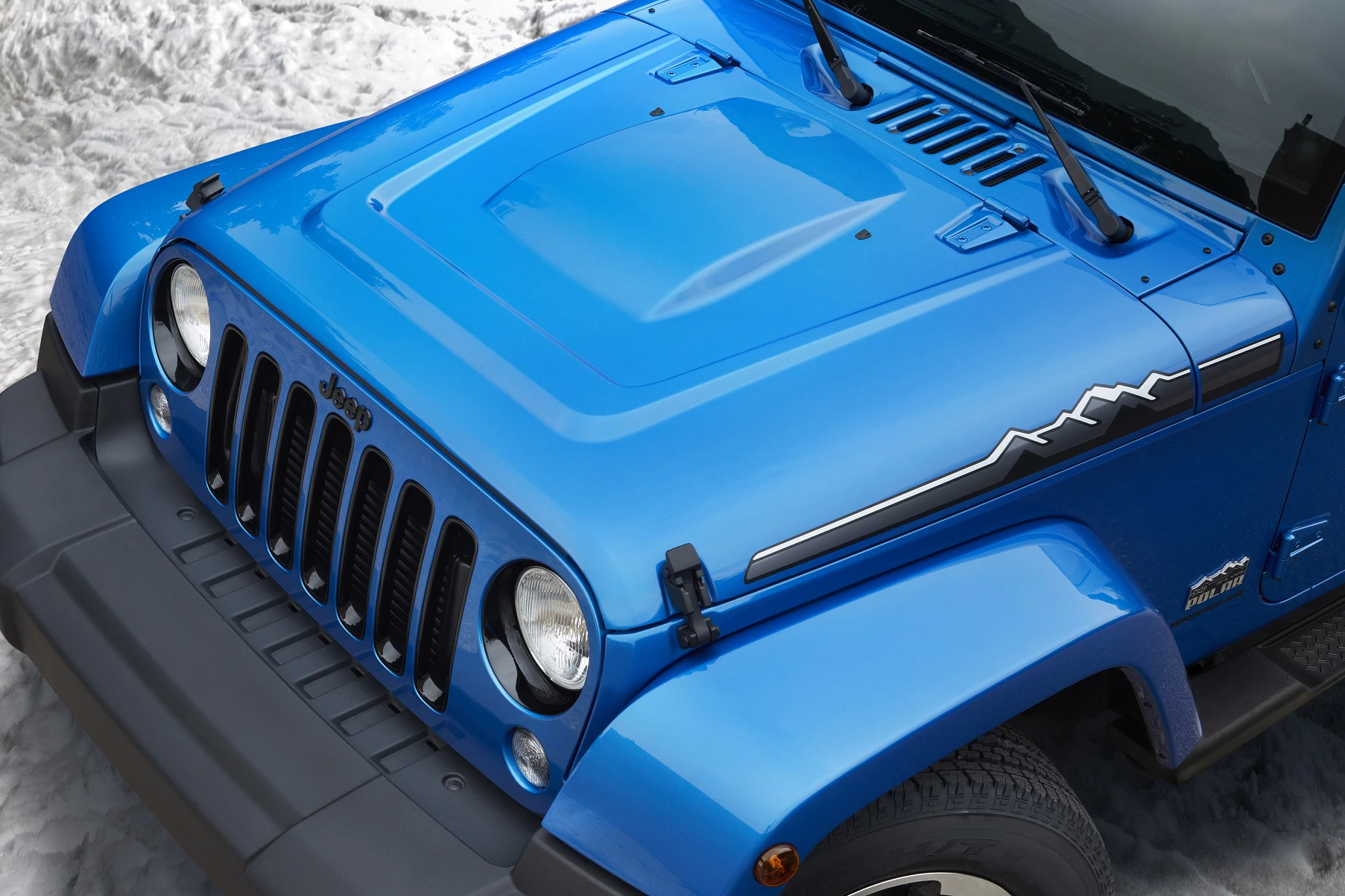 Blauer Jeep Wrangler mit Bergmotiv auf der Motorhaube, abgestellt auf schneebedecktem Boden. Vorderansicht zeigt markantes Kühlergrilldesign.