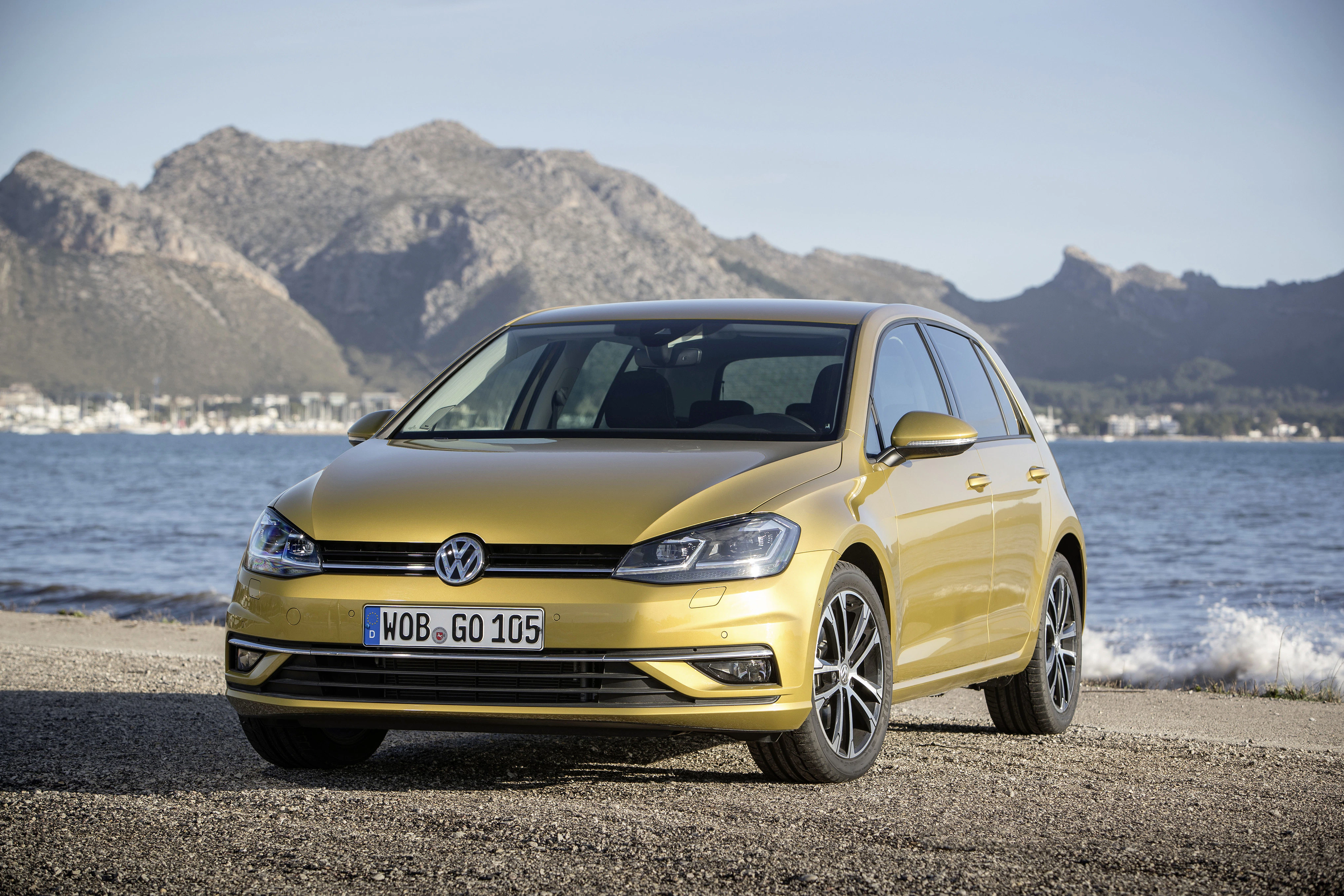 Goldener VW Golf steht an einem Strand mit Bergen im Hintergrund bei sonnigem Wetter.