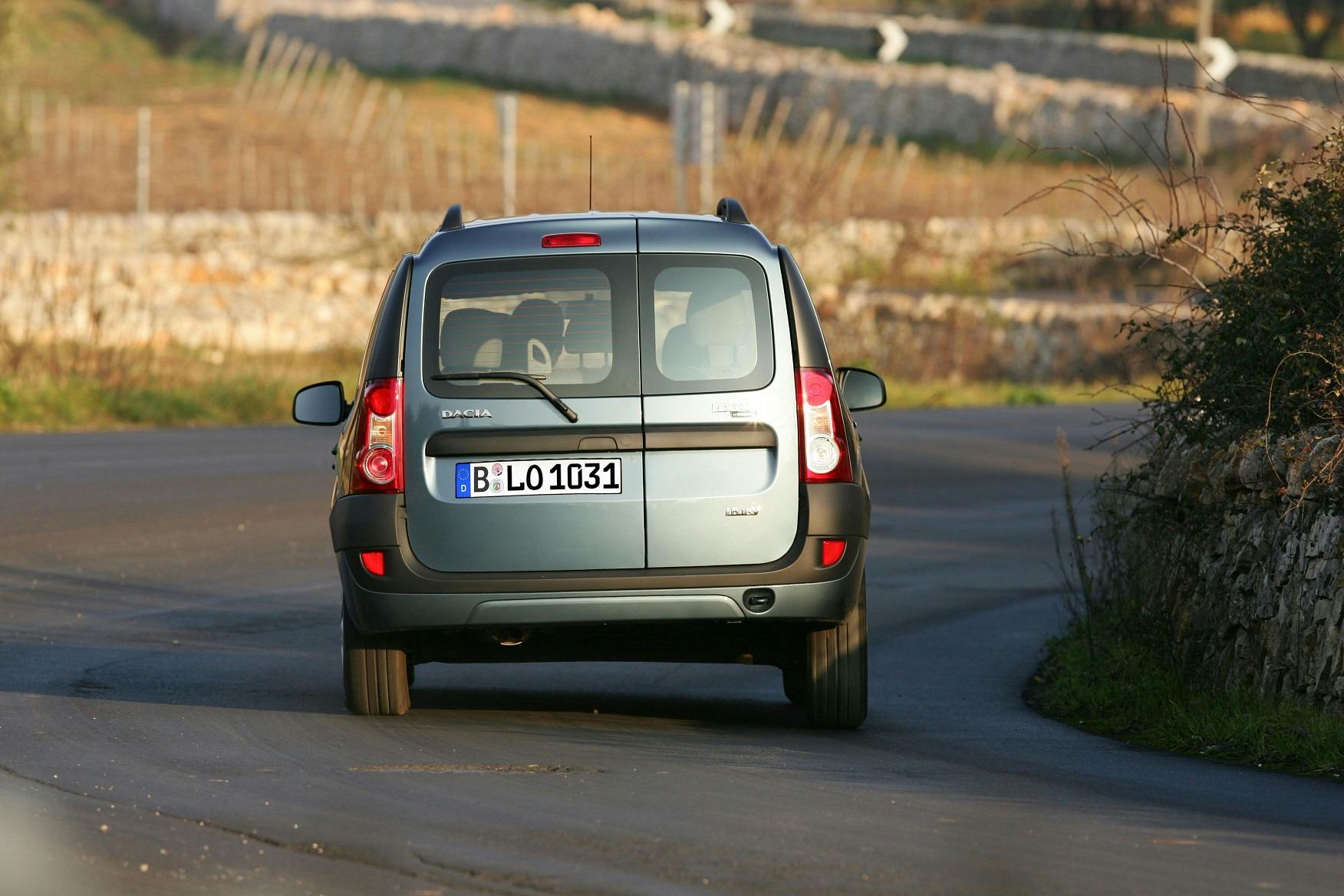 Ein grauer Dacia Logan MCV fährt auf einer ländlichen, kurvigen Straße. Das Auto hat ein deutsches Kennzeichen.