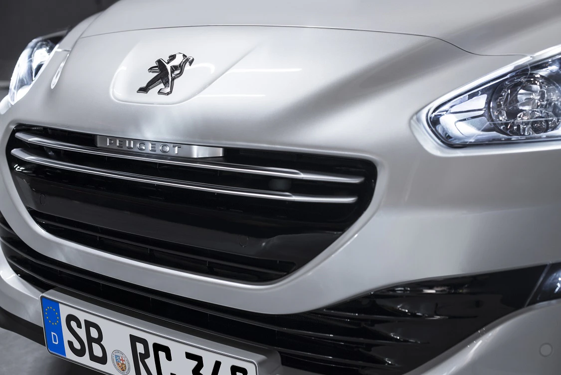 Nahaufnahme der Front eines silbernen Peugeot-Autos mit leuchtendem Scheinwerfer und sichtbarem Nummernschild.