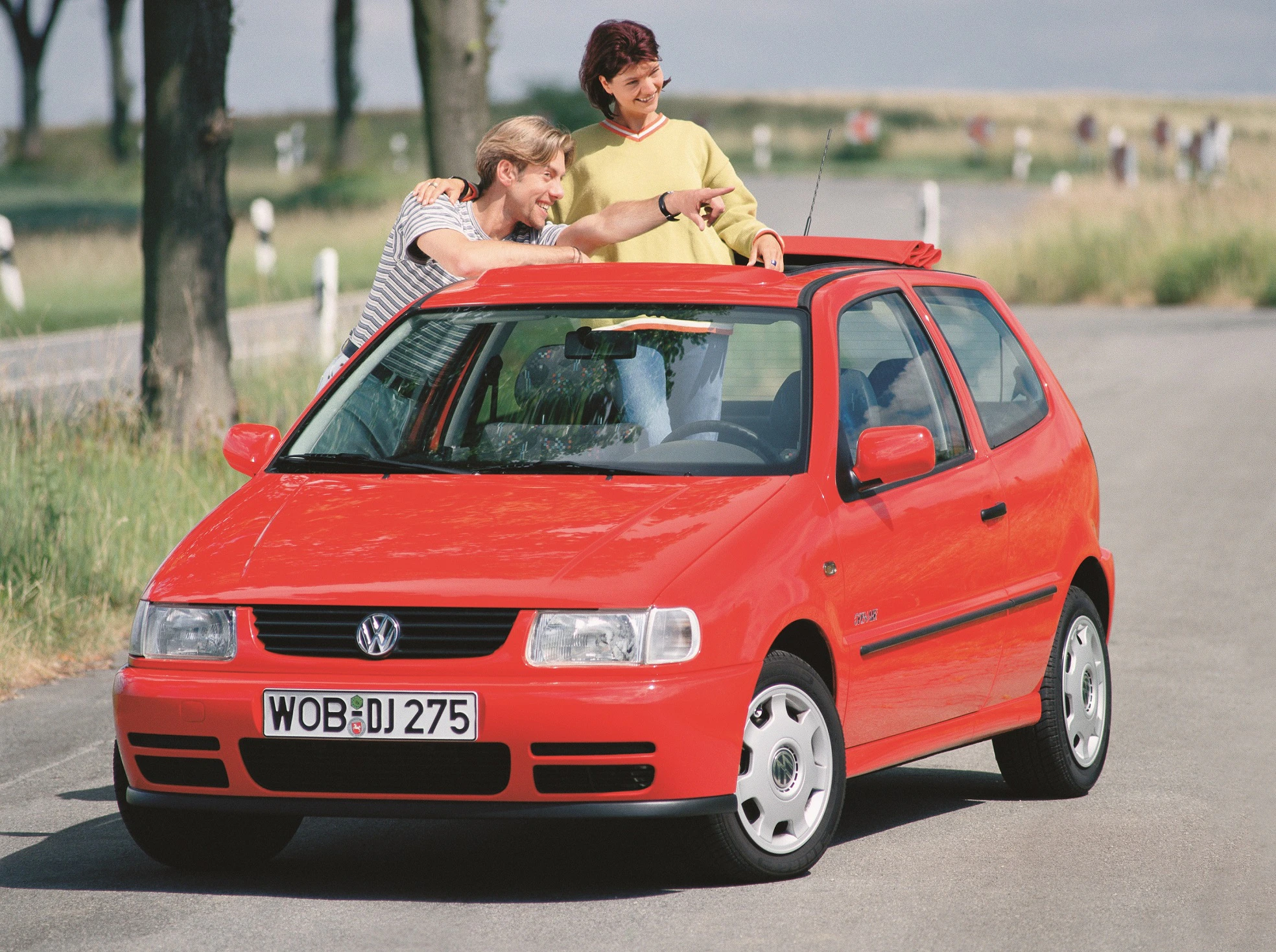 Ein roter VW Polo steht auf einer Landstraße. Ein Mann und eine Frau stehen aus dem Schiebedach und zeigen auf etwas außerhalb des Bildes.
