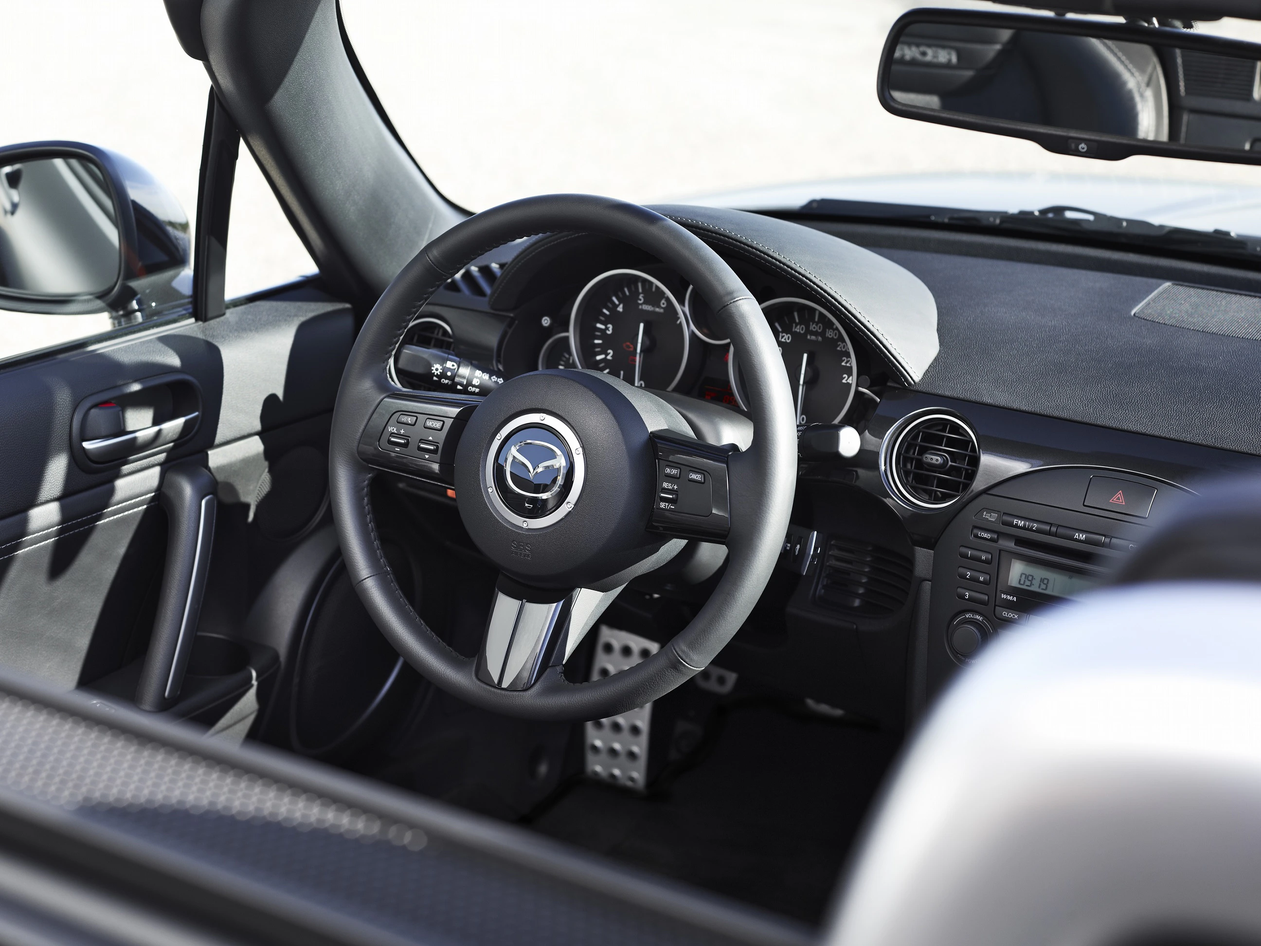 Innenraum eines Autos mit schwarzem Lenkrad und Mazda-Logo. Sicht auf Armaturenbrett, Tachometer und Pedale. Sportsitze teilweise sichtbar.