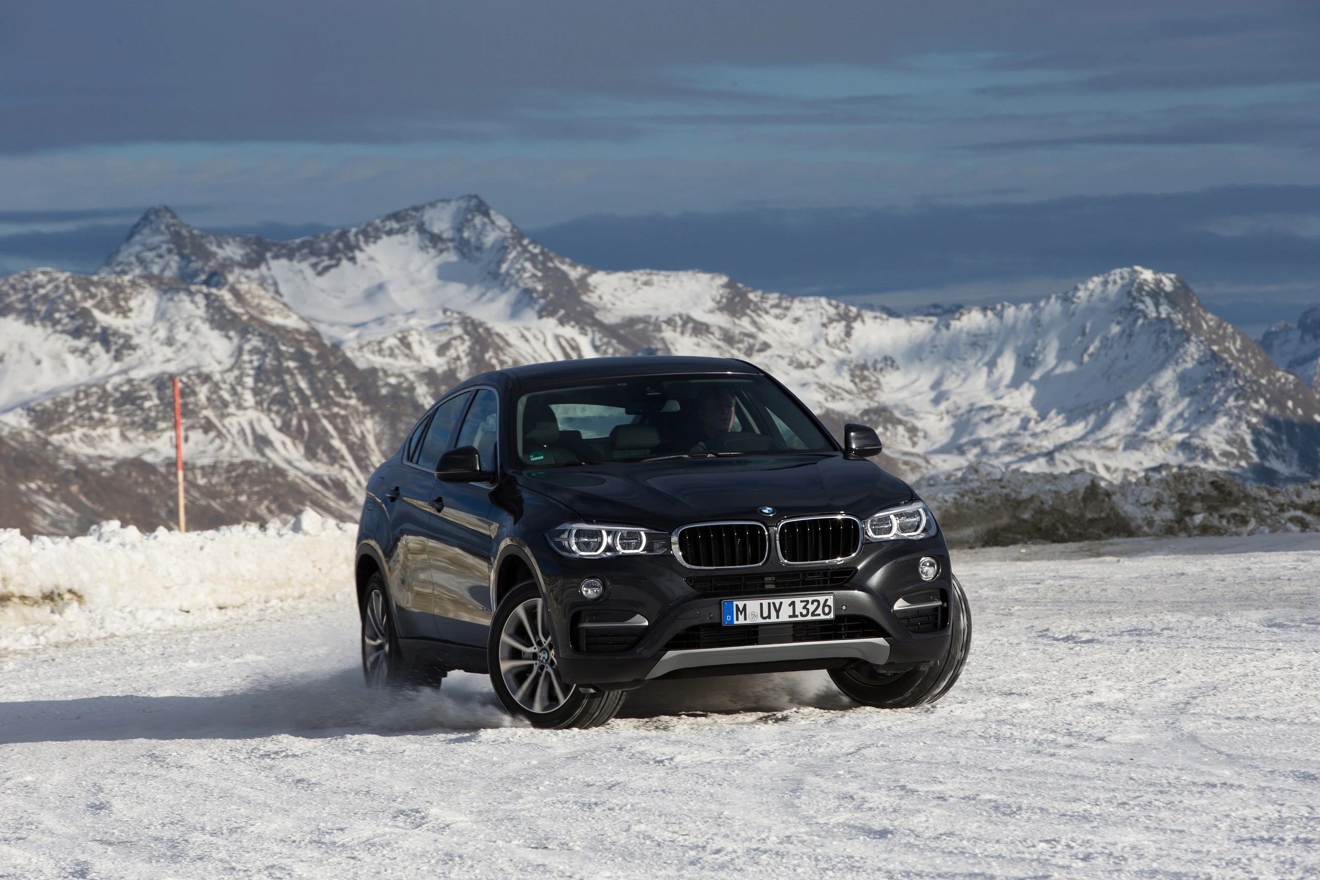 Schwarzer SUV fährt auf schneebedeckter Straße in den Bergen. Majestätische schneebedeckte Gipfel im Hintergrund unter bewölktem Himmel.