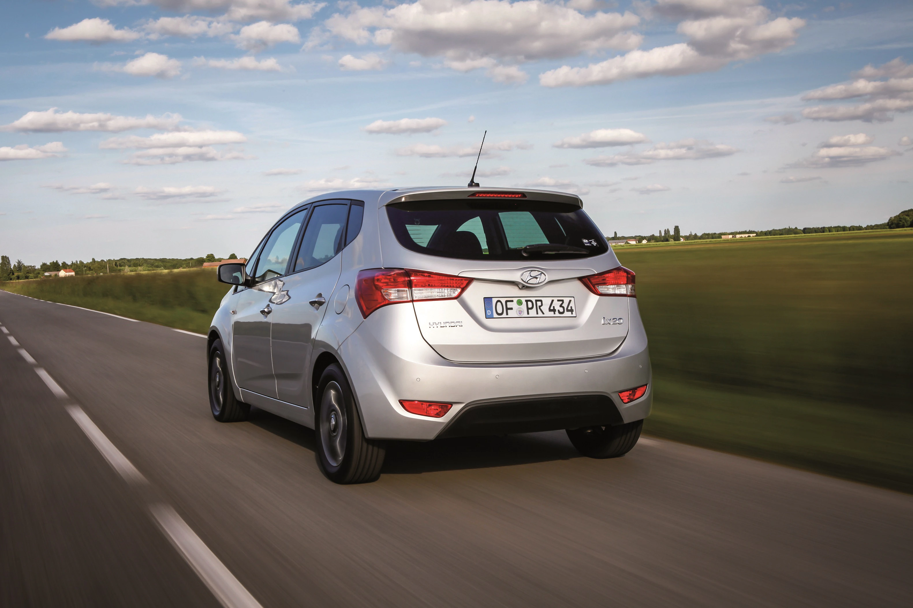 Silberner Hyundai i20 fährt auf einer Landstraße, umgeben von grünen Feldern und einem wolkigen Himmel.