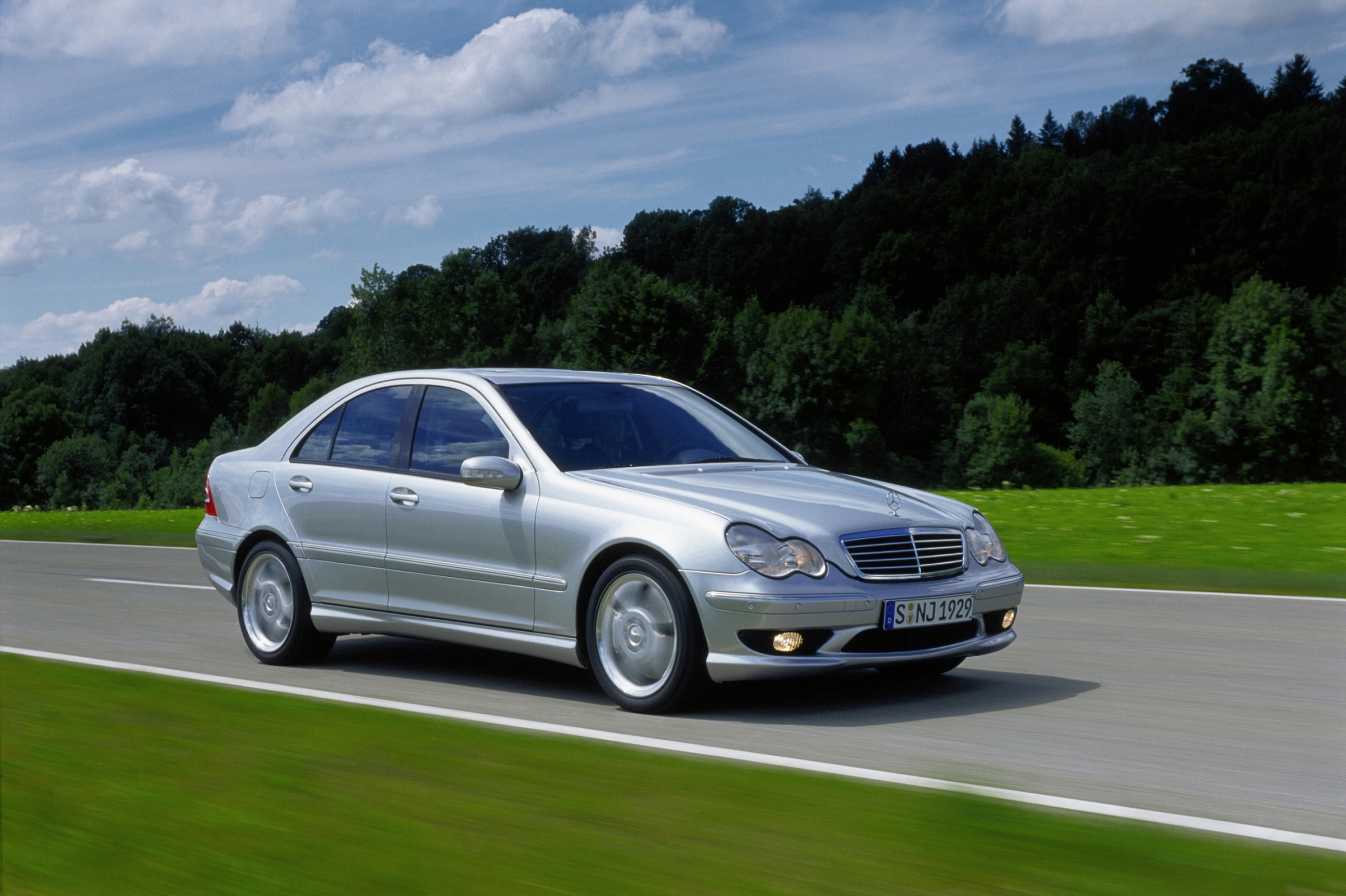 Silberner Mercedes-Limousine fährt auf einer Landstraße bei sonnigem Wetter, umgeben von grüner Landschaft und Bäumen im Hintergrund.