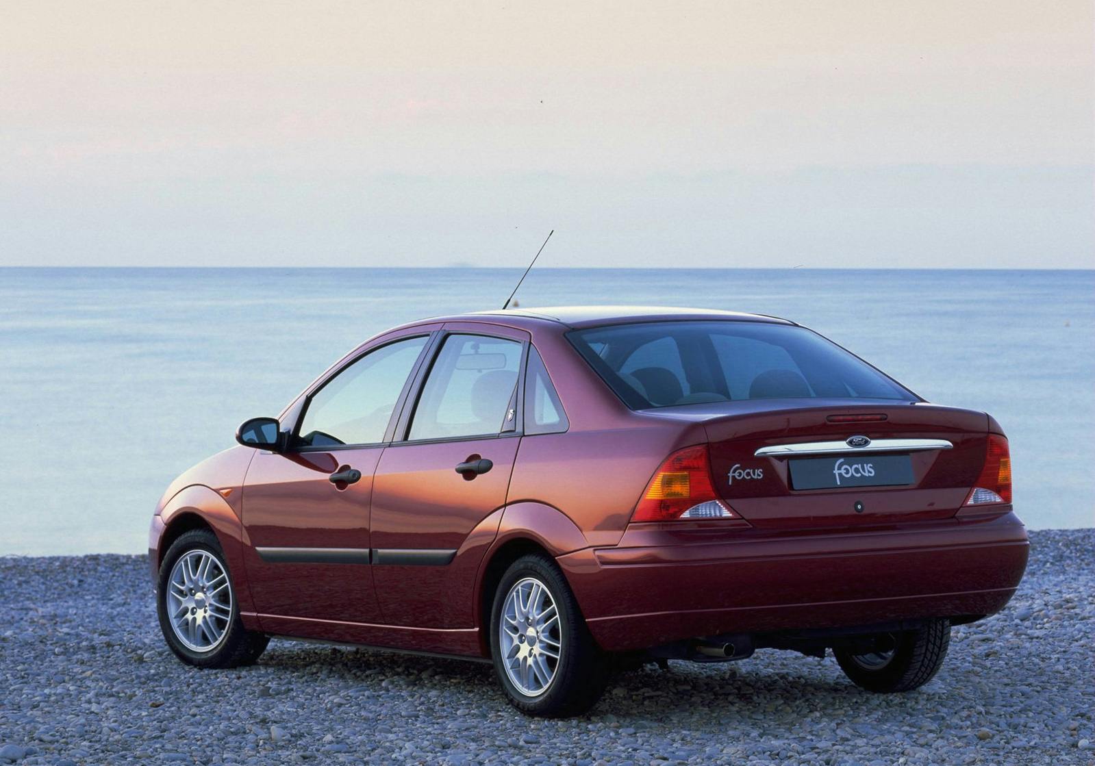 Roter Ford Focus parkt auf einem kiesigen Strand mit Blick auf das ruhige Meer und den pastellfarbenen Himmel im Hintergrund.