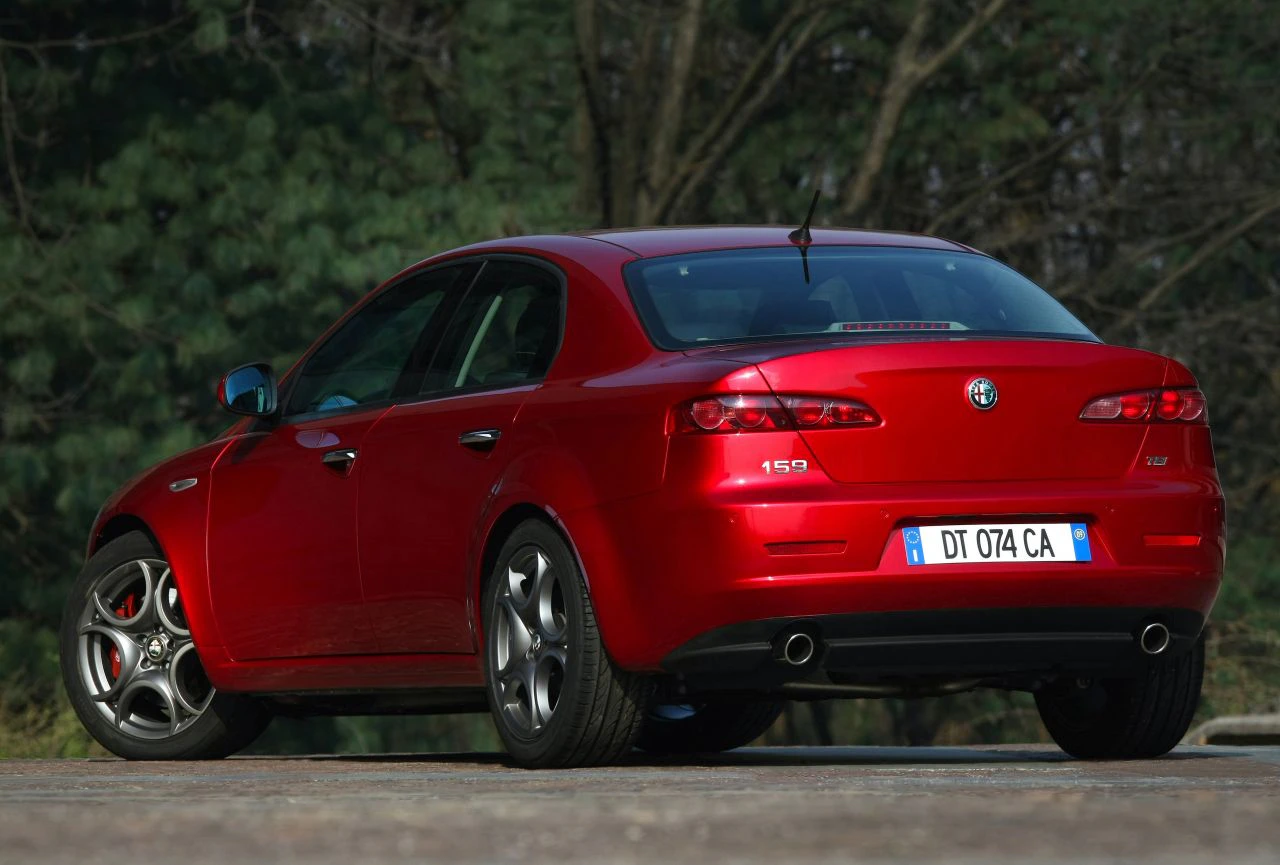 Roter Alfa Romeo 159 in Seiten- und Heckansicht, geparkt auf einer Straße vor einem Hintergrund mit Bäumen.