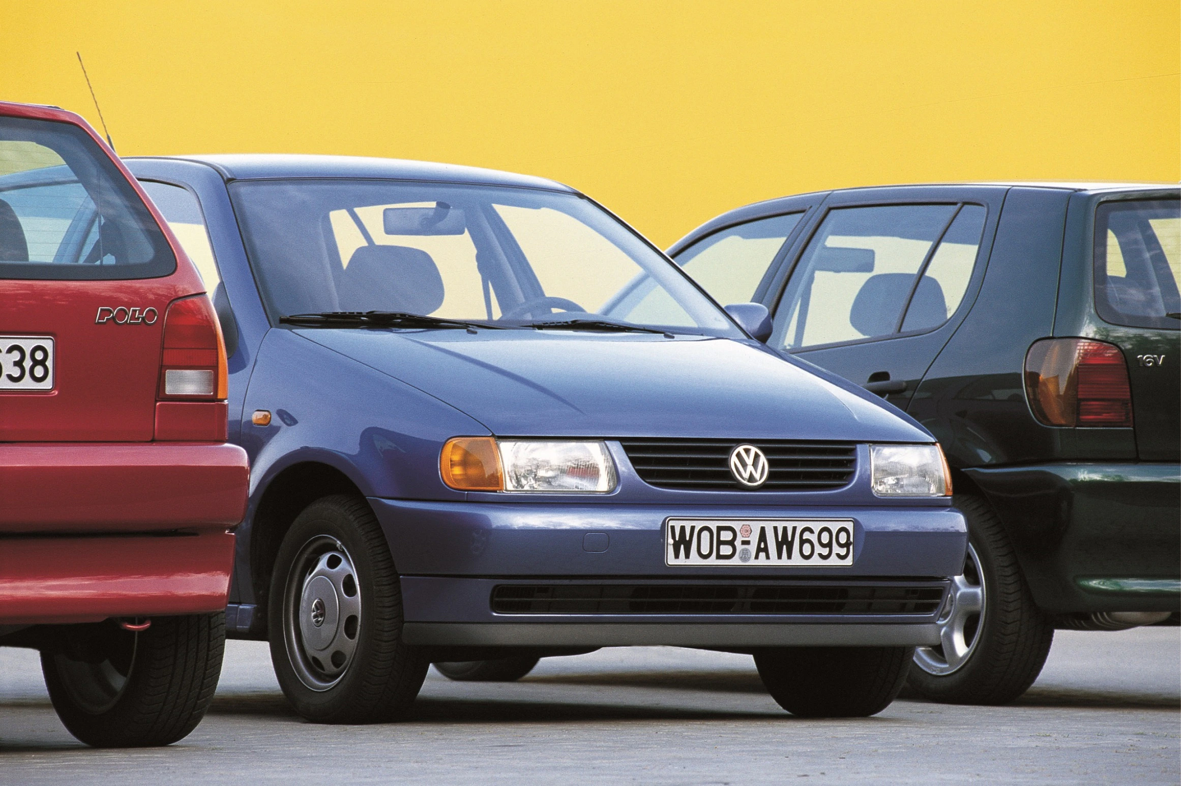 Ein blauer Volkswagen Polo ist zwischen zwei Autos geparkt, eines rot, das andere schwarz, vor einer gelben Wand.