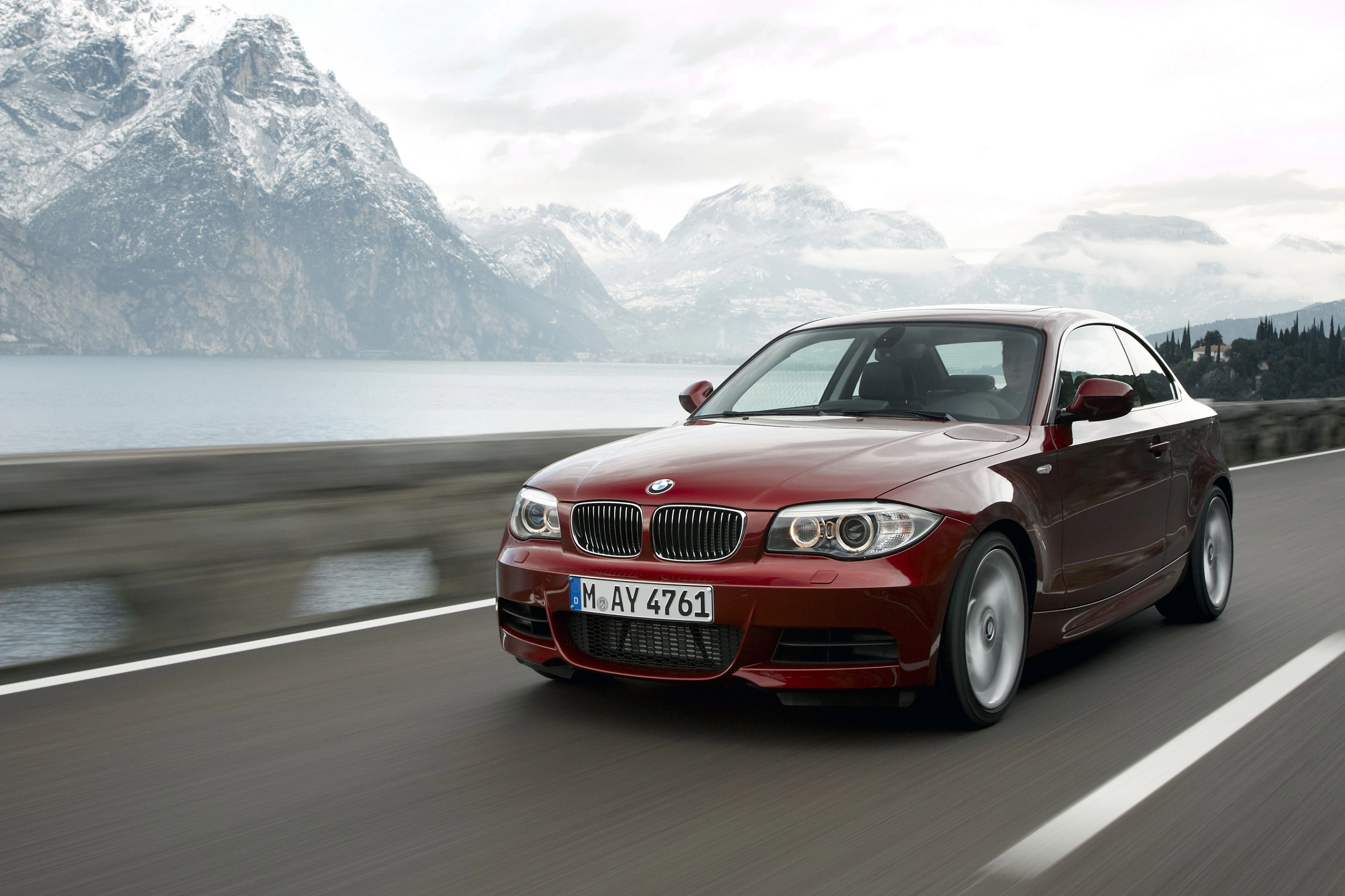 Rotes BMW-Coupé fährt auf einer Straße entlang eines Sees, umgeben von schneebedeckten Bergen unter bewölktem Himmel.
